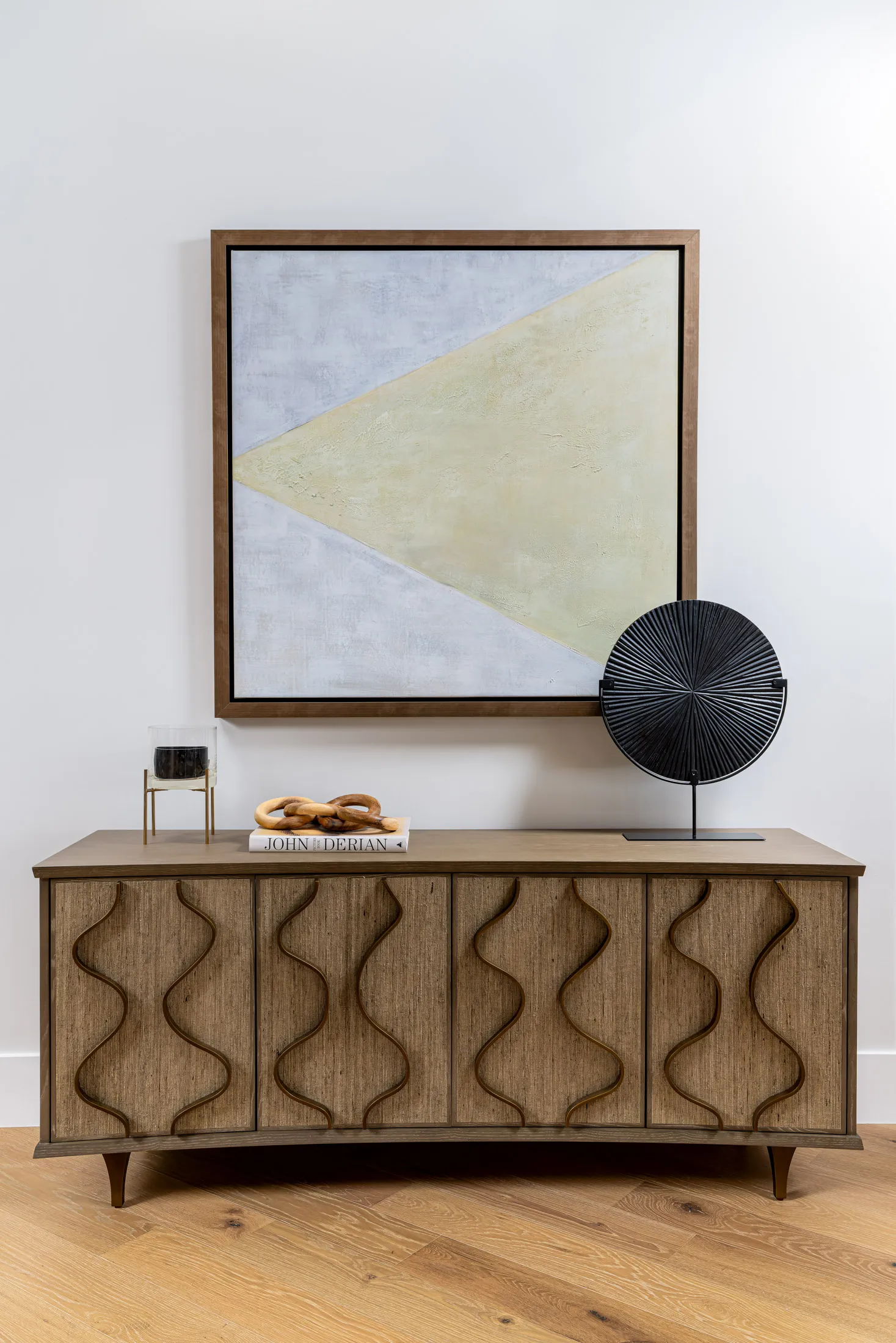 A textured wooden console with curved front panels sits beneath a large geometric painting, displaying a black disc sculpture and simple decor.