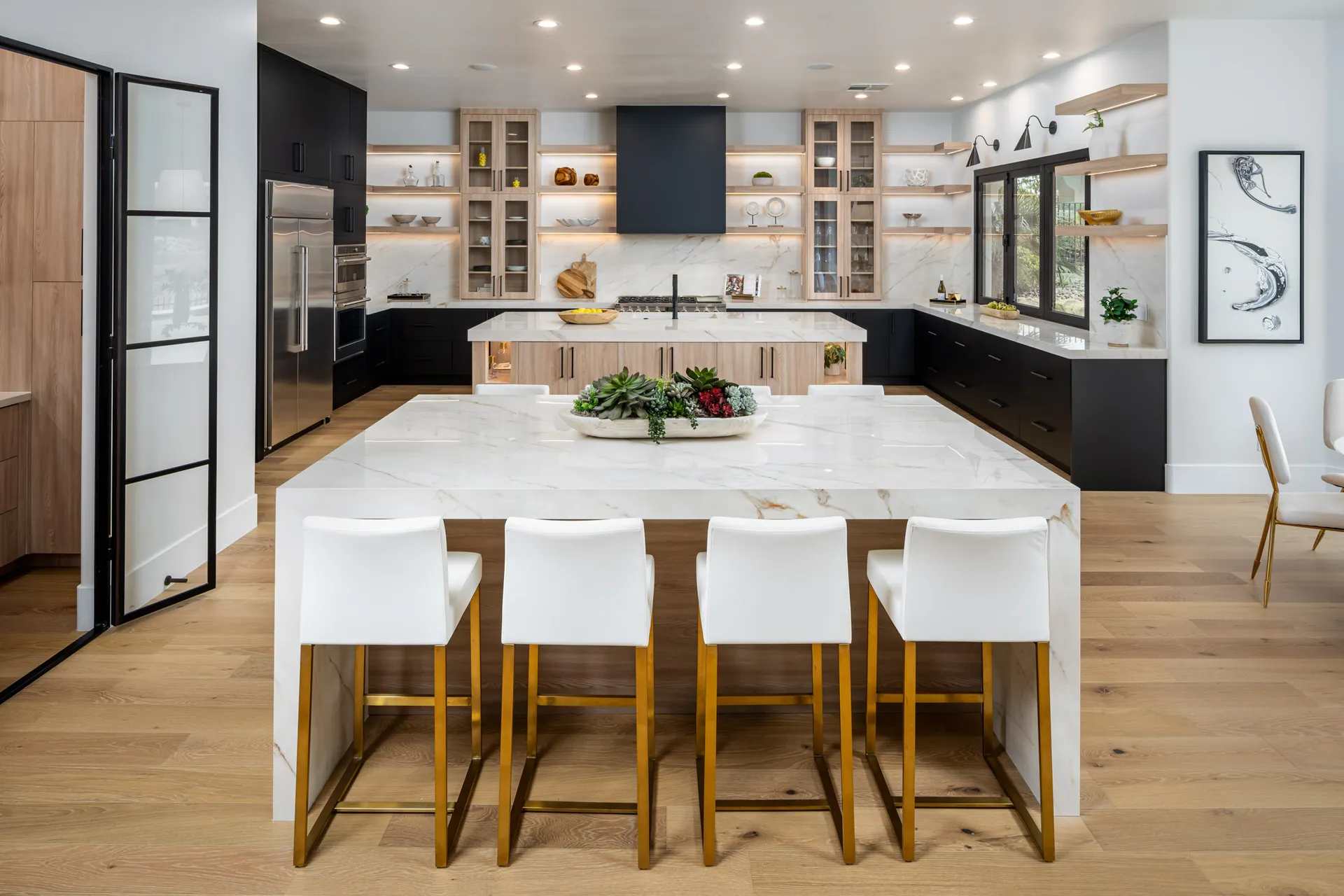 A modern kitchen with a large marble-topped island, wood and black cabinetry, and white barstools featuring gold legs.
