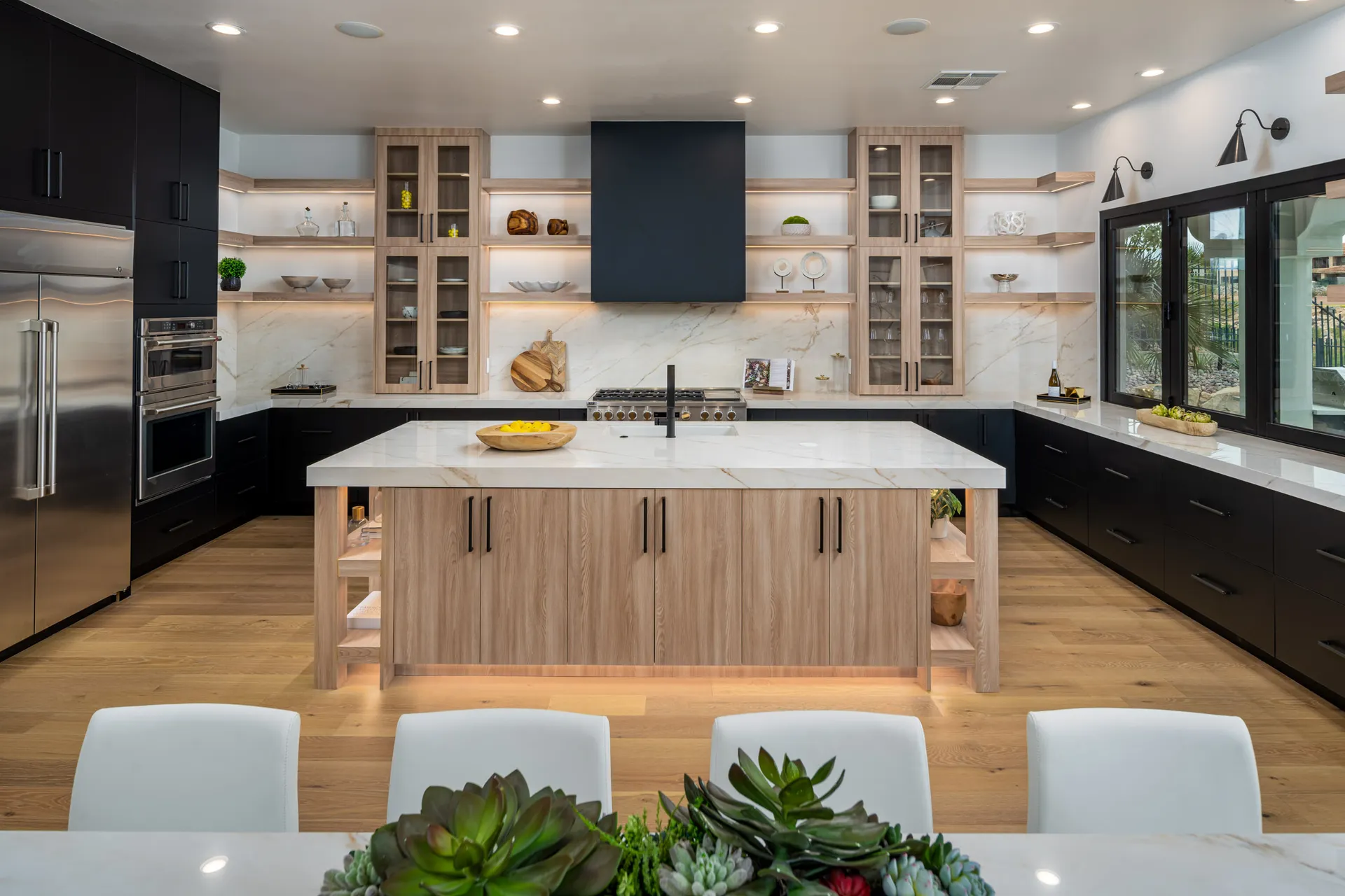 A spacious kitchen with black lower cabinets, light wood cabinetry, and a large marble-topped island with integrated shelving.