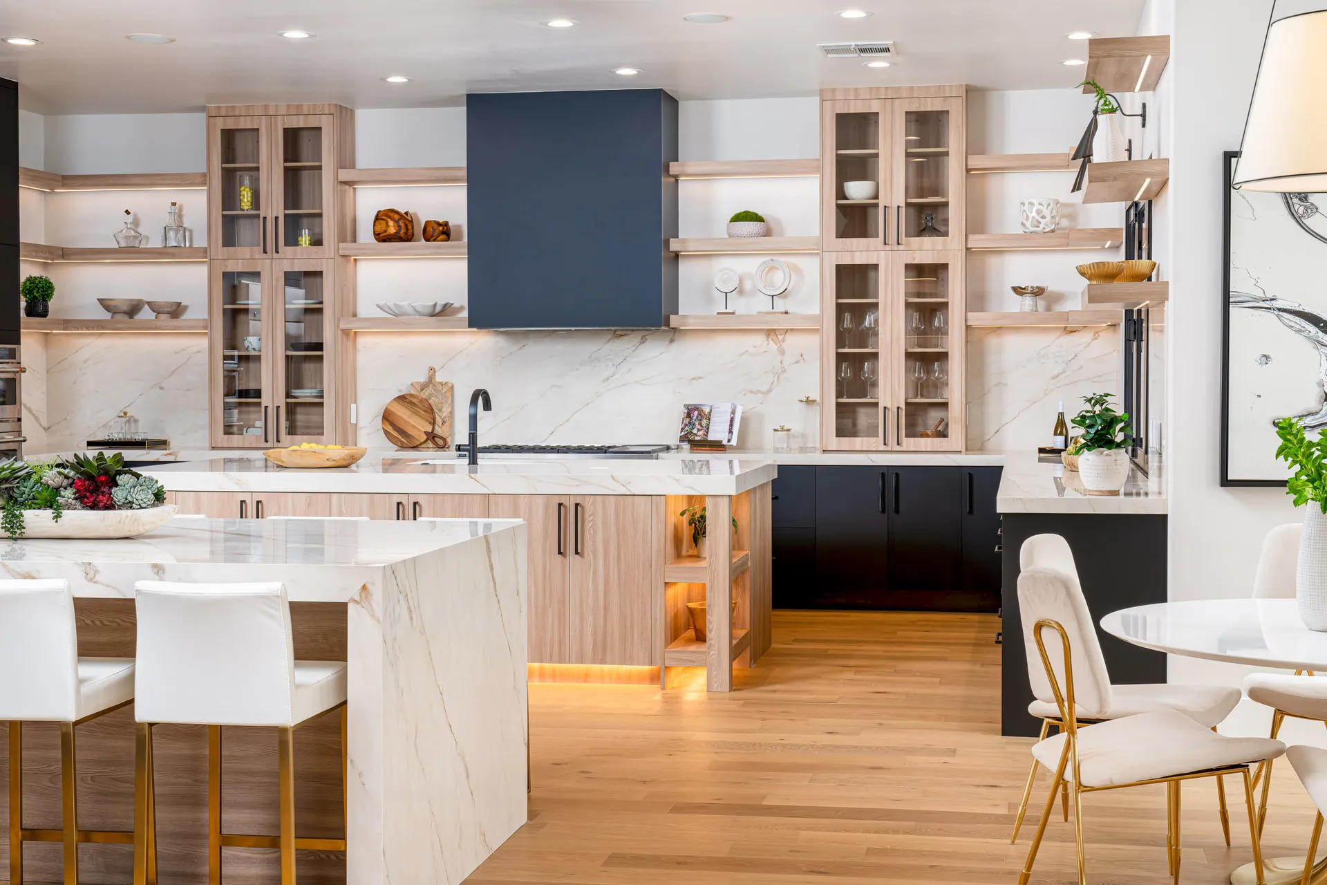 A contemporary kitchen with marble countertops, light wood cabinets, and open shelves, centered around a large island.
