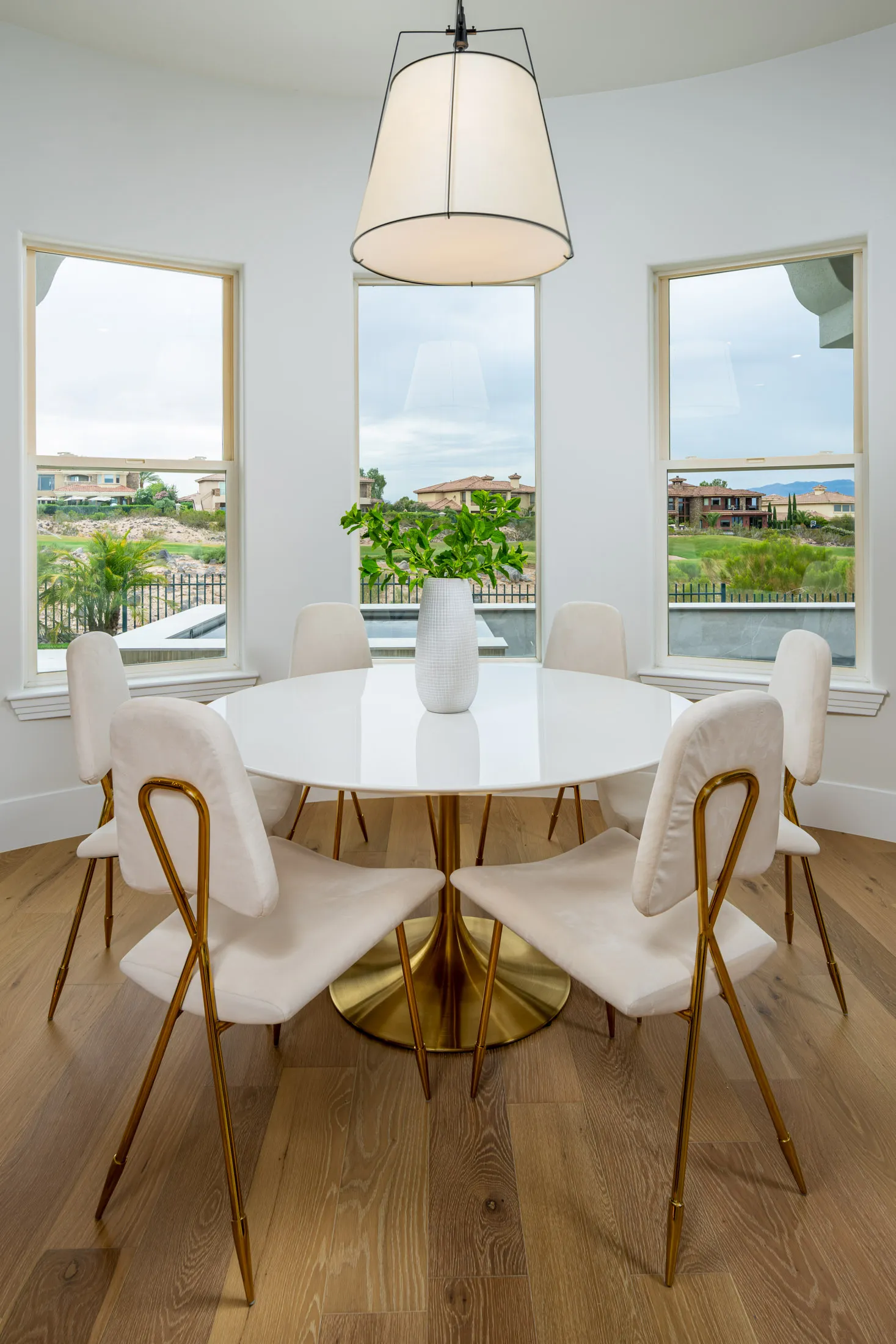 A round white table with a gold base is surrounded by white chairs with gold legs, all beneath a large pendant light.