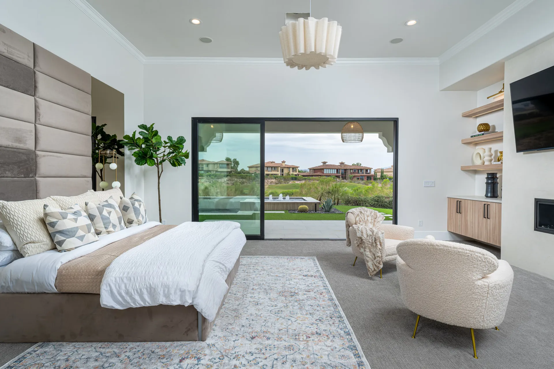 A spacious bedroom with a tall tufted headboard, plush chairs, and sliding glass doors revealing an outdoor fountain view.