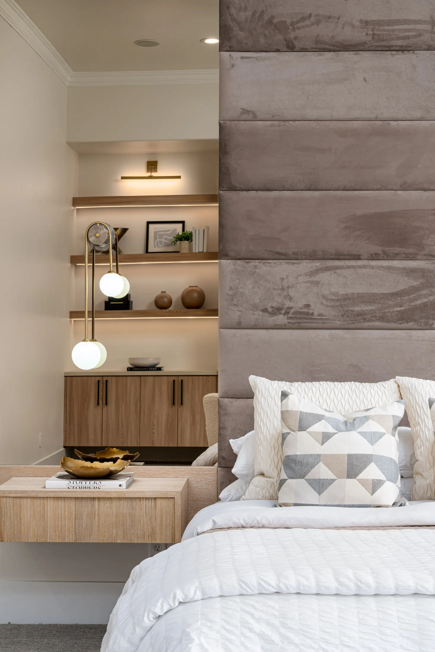 A tall upholstered headboard in soft gray beside built-in wooden shelves with hanging globe lights.