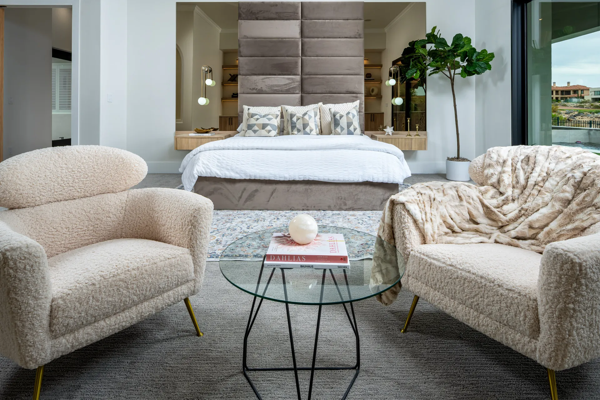 A bedroom with a tall upholstered headboard, two plush chairs, and a glass coffee table in front.