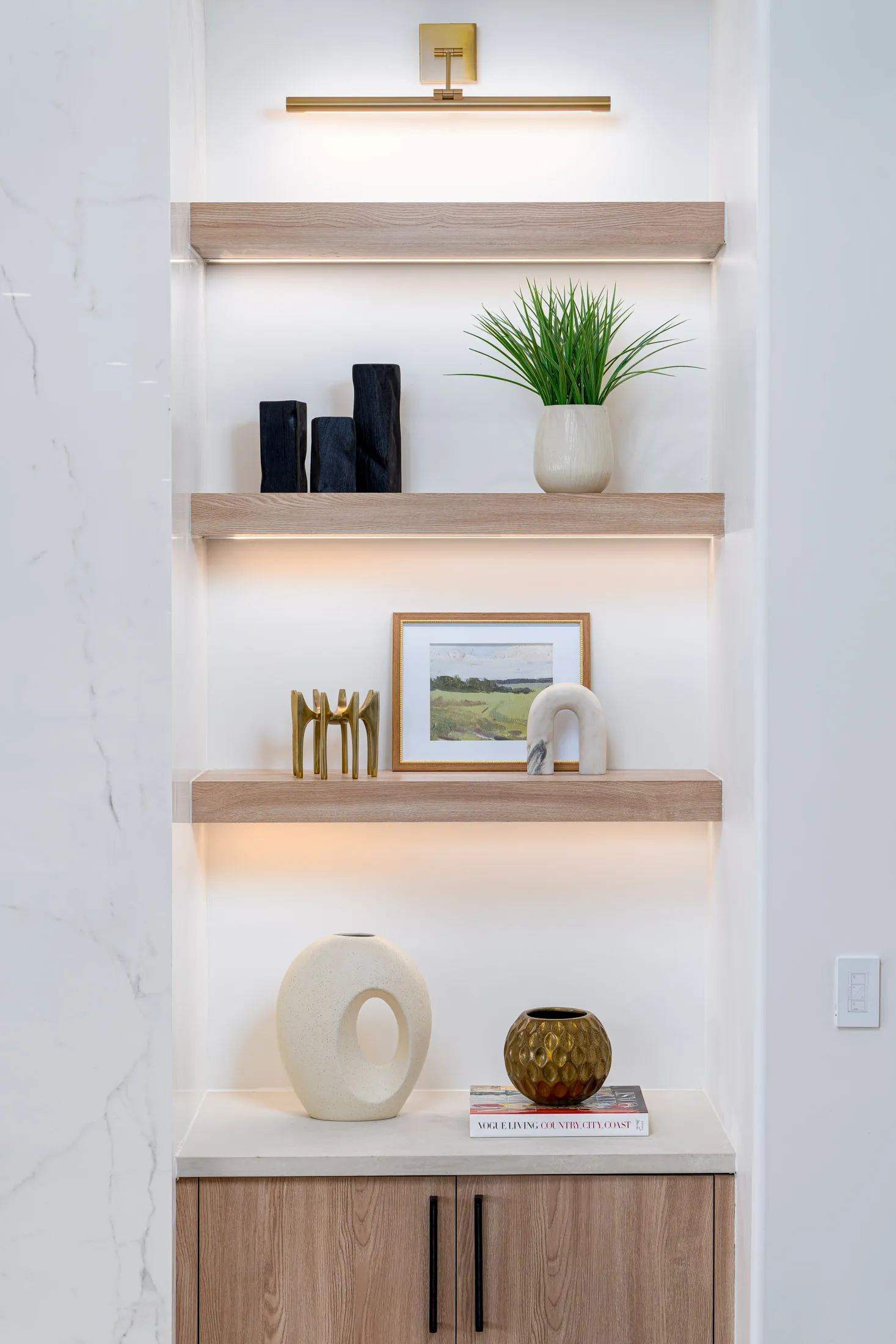 Wooden floating shelves featuring sculptural pieces, a potted plant, and a framed landscape, illuminated by a brass picture light.
