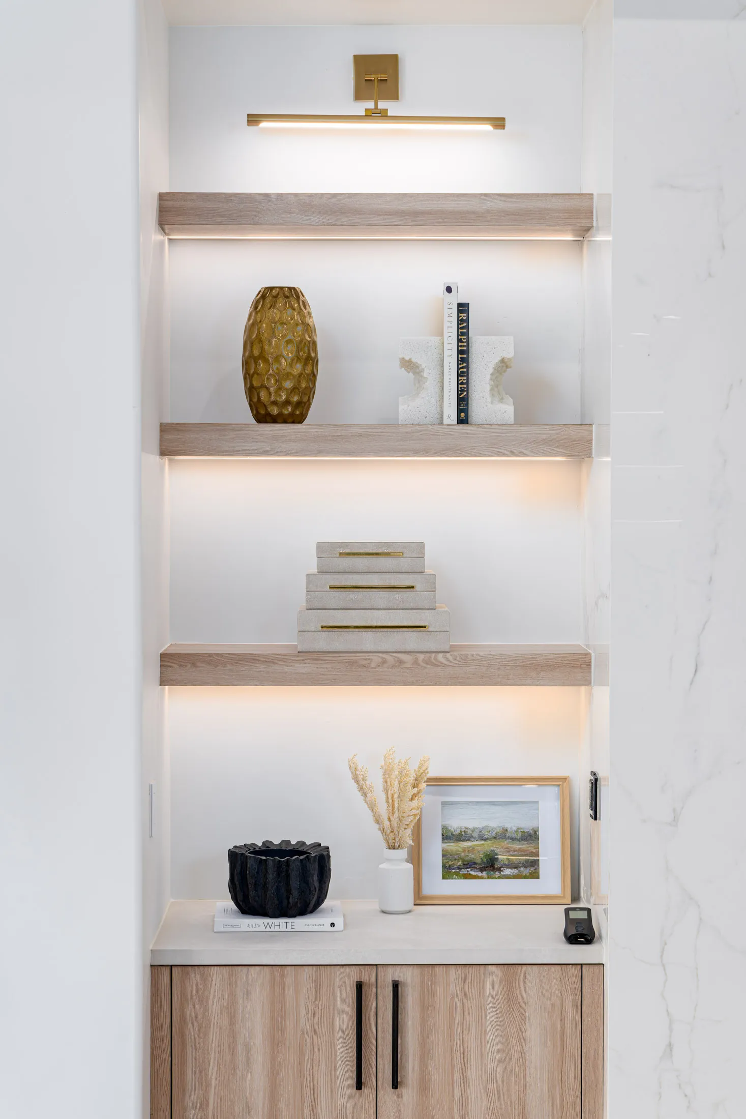 Wooden floating shelves with integrated lighting displaying a gold vase, decorative bookends, and boxes, above a cabinet with a small vase and framed art.