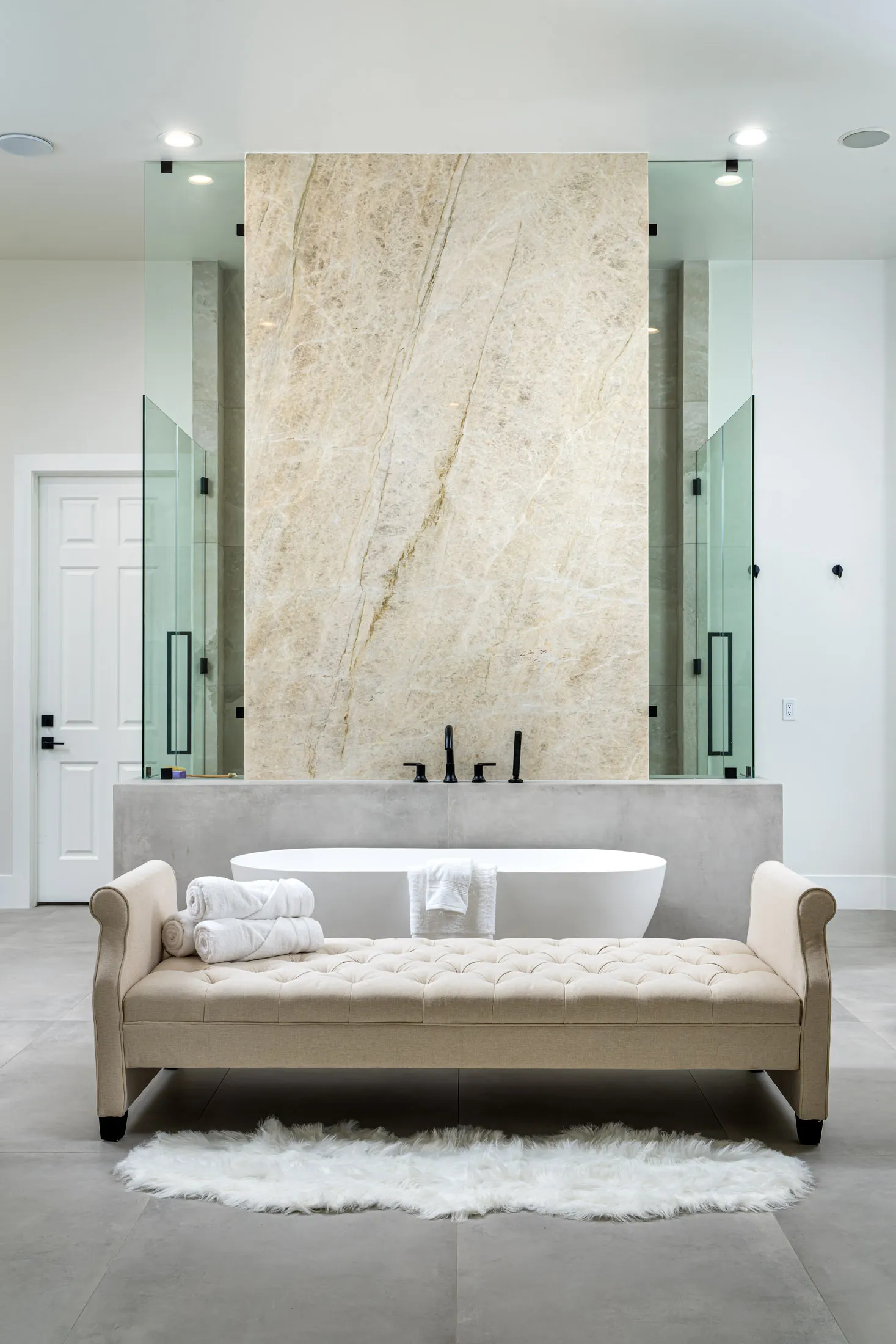 A freestanding white bathtub set against a tall marble-look wall, with a tufted bench in front.