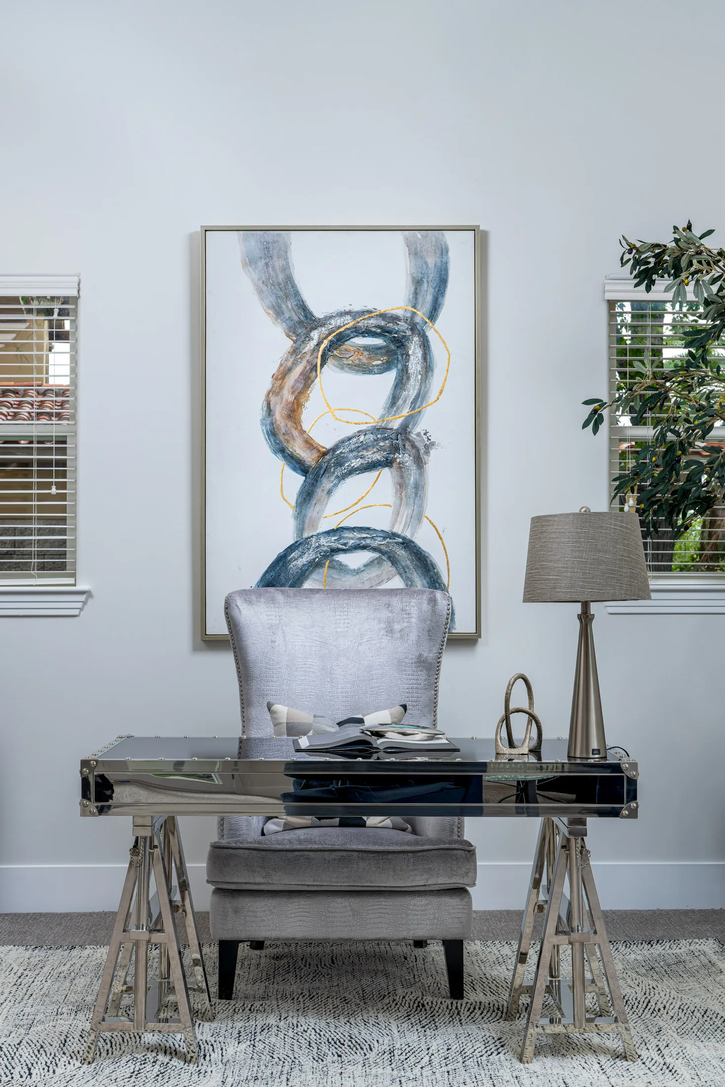 A reflective metal desk paired with a plush gray chair in front of a large abstract chain painting on the wall.
