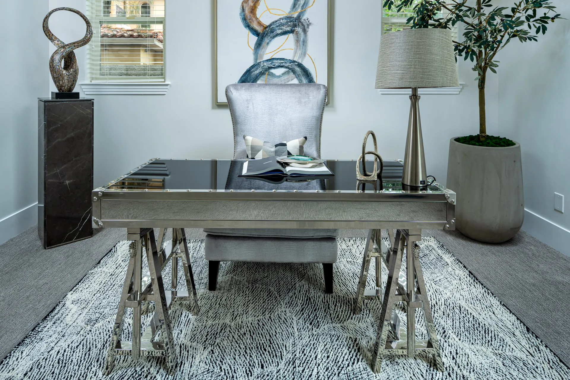 A polished metallic desk on trestle stands, paired with a gray accent chair and abstract artwork. A tall lamp and sculptural pieces complete the modern look.