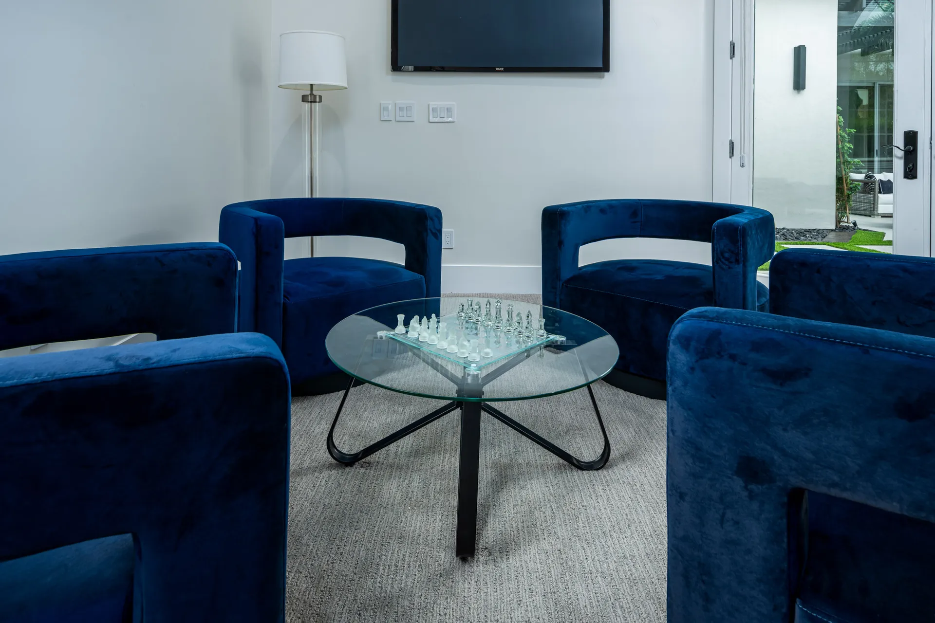 Four round blue velvet chairs arranged around a circular glass coffee table with a clear chess set. A floor lamp and a wall-mounted TV are seen in the background.