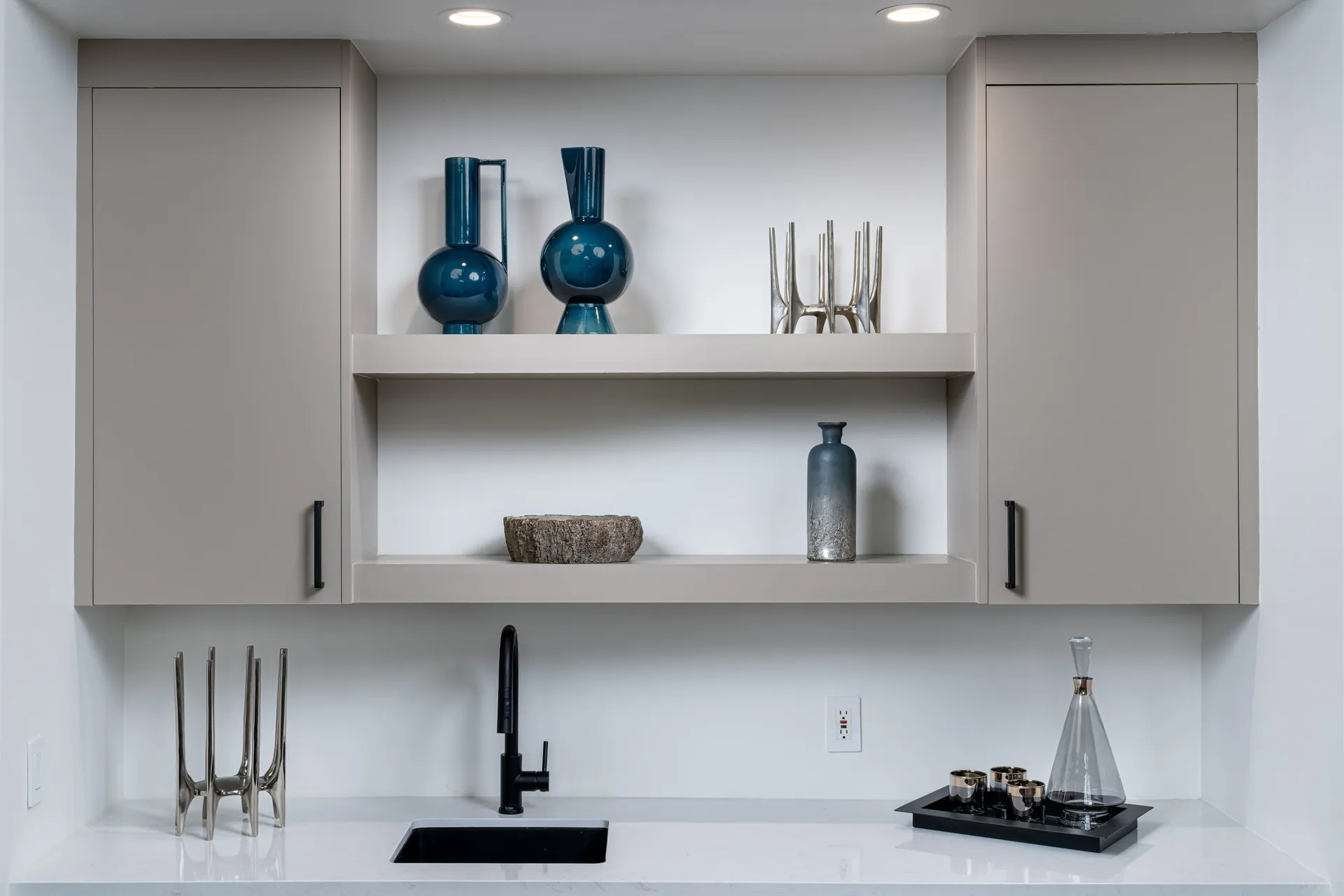 A built-in wet bar with neutral cabinets, open shelving, and a black faucet. Blue ceramic vases and metallic sculptures add decorative flair.