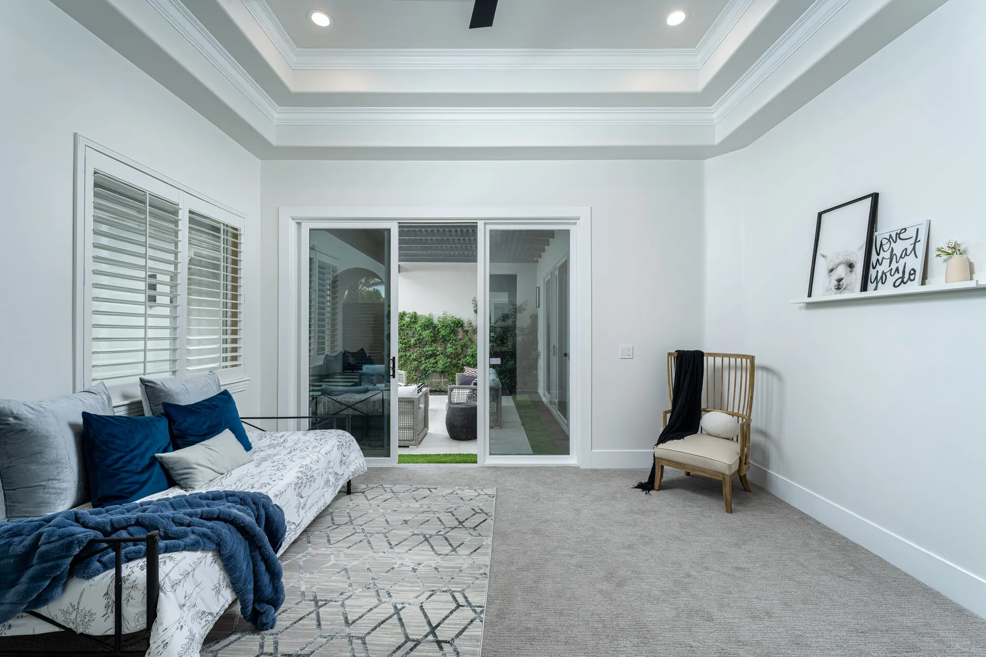 A bright sitting area featuring a daybed with layered pillows and sliding doors leading to an outdoor seating space. A wooden accent chair and simple artwork complete the room.