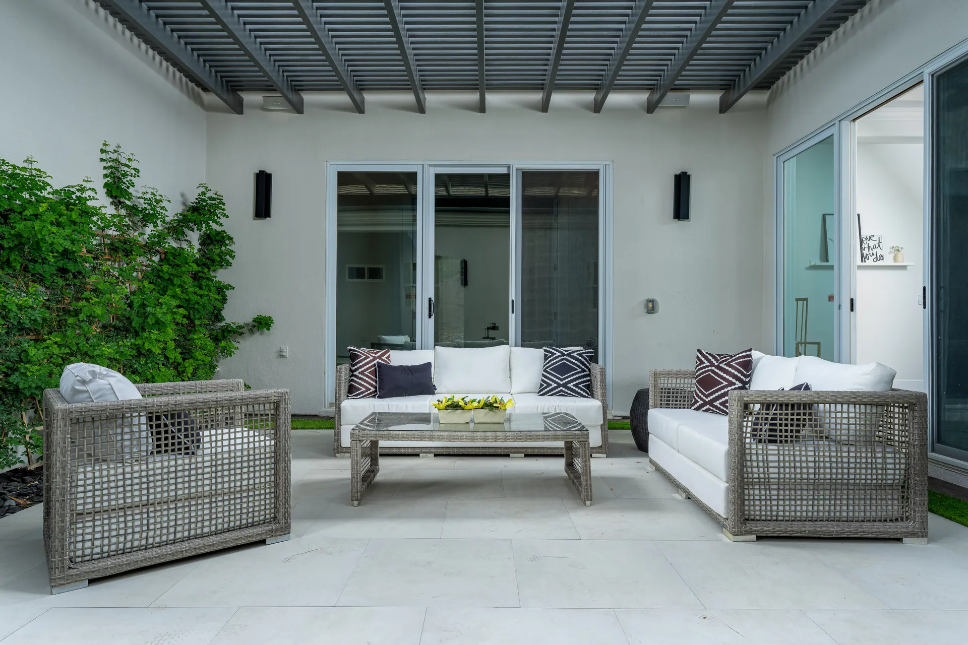 A covered patio area with woven rattan-style seating, white cushions, and geometric accent pillows. Greenery lines one wall, and glass doors lead indoors.