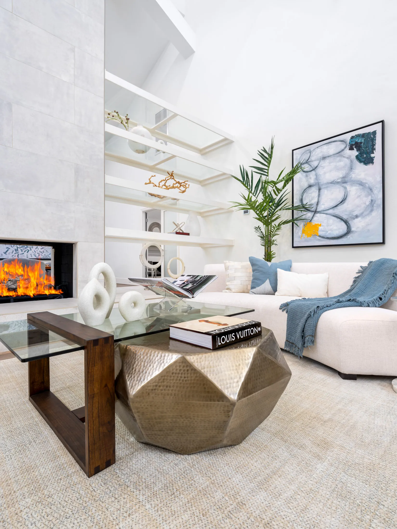 A modern living room with a hammered metallic coffee table, a layered glass top, and a lit fireplace near floating white shelves.