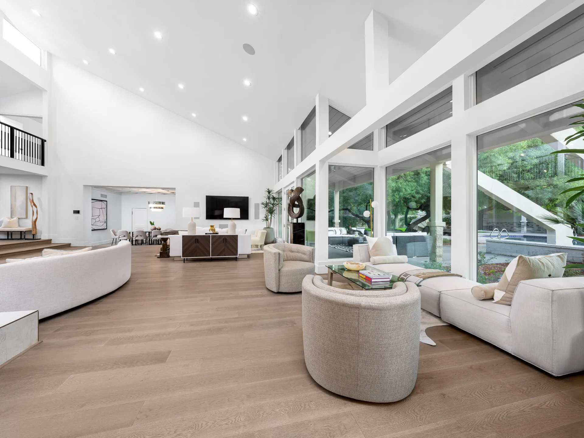 A bright, open living area with tall ceilings, curved white seating, and expansive windows overlooking a patio and pool.