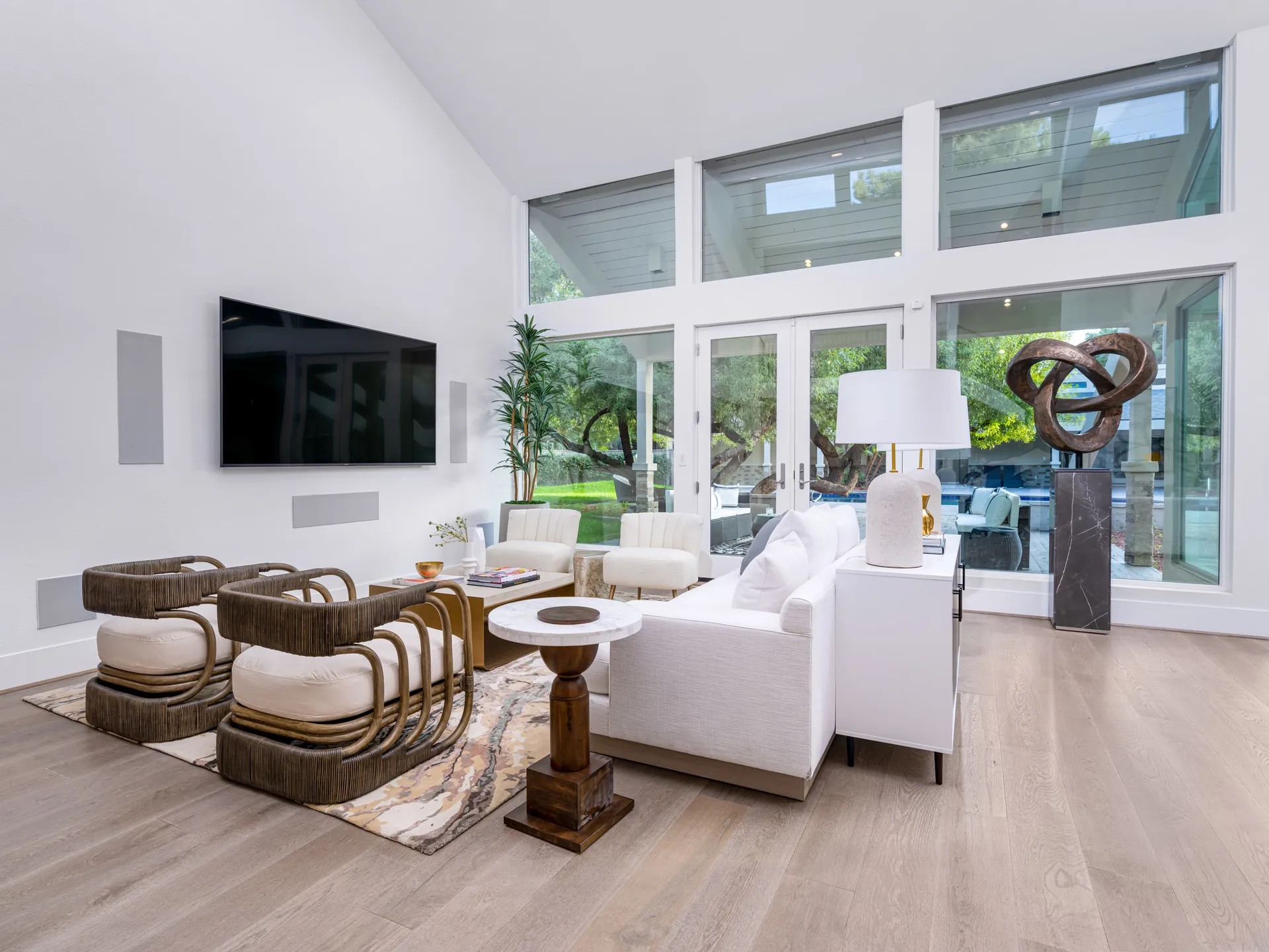 A bright living room with two sculptural rattan chairs on a patterned rug, a white sofa, and glass doors opening to a patio and pool area.
