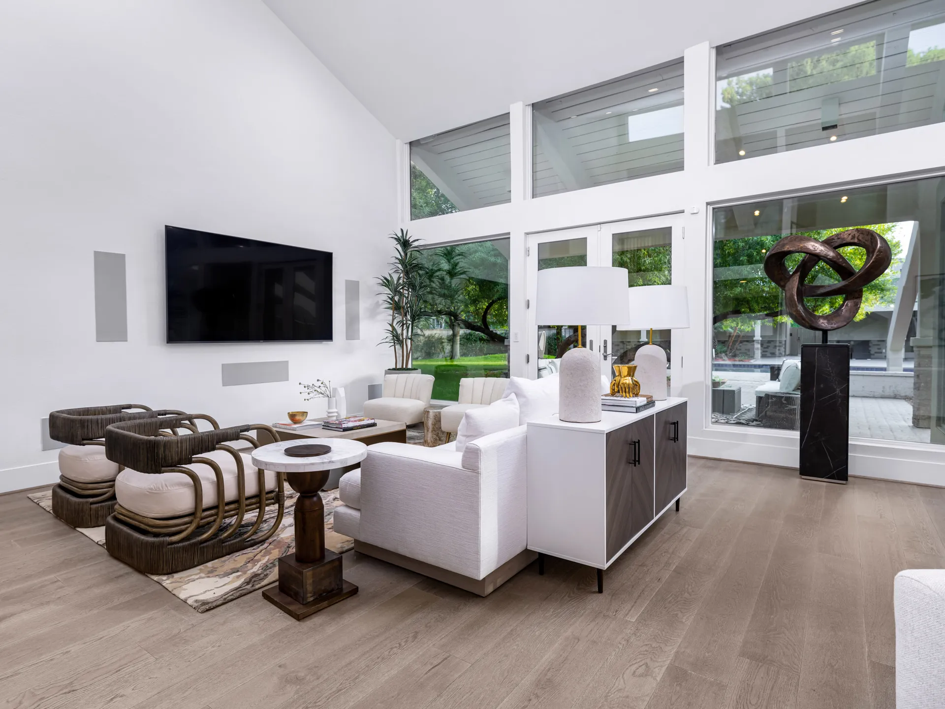 A spacious living room featuring two rattan-frame chairs with plush cushions, a wall-mounted TV, a white-and-wood console, and glass doors overlooking the yard.
