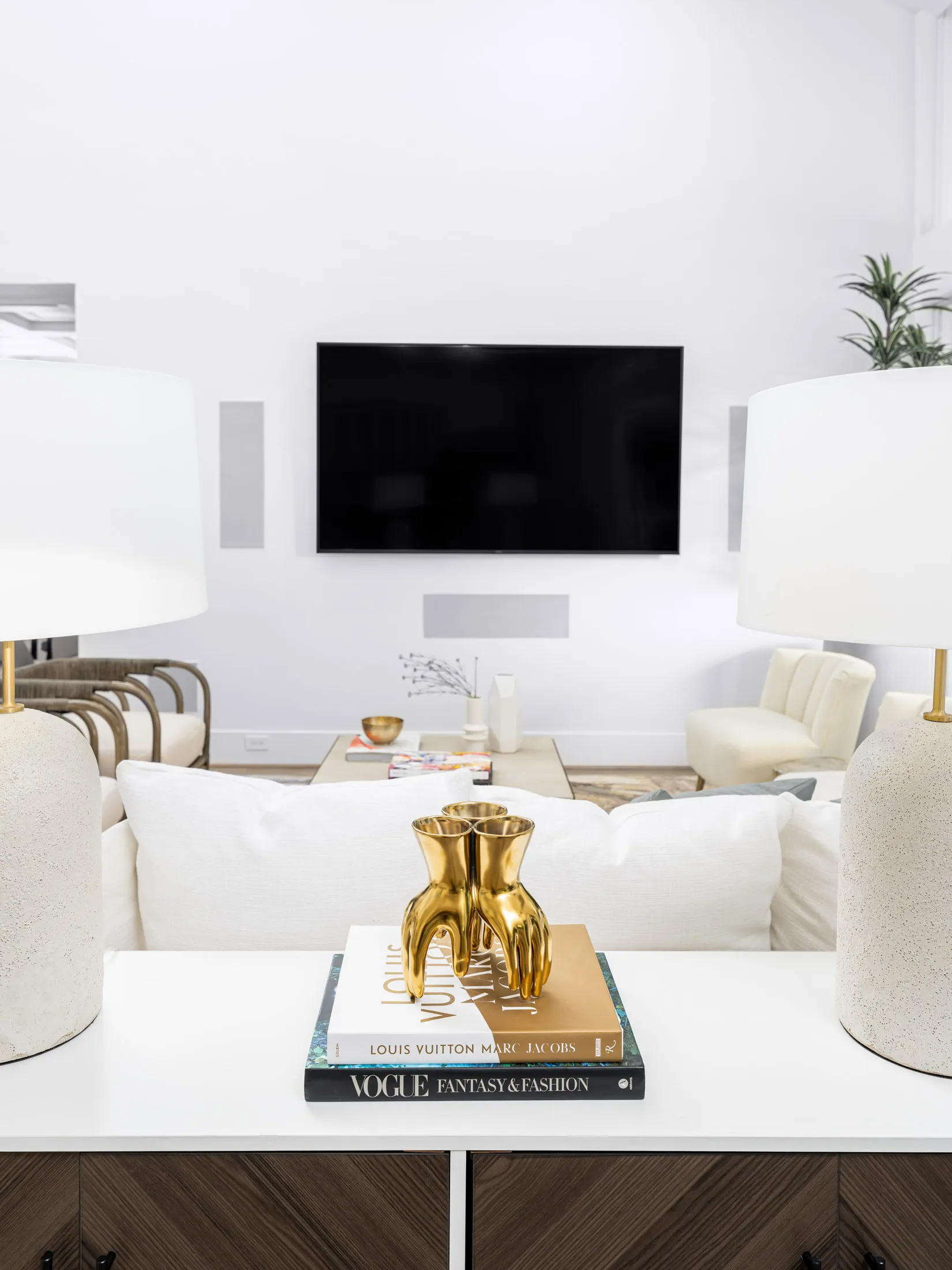 A white console table behind a sofa, featuring a gold hand sculpture atop fashion books, with a large TV mounted on the wall in the background.
