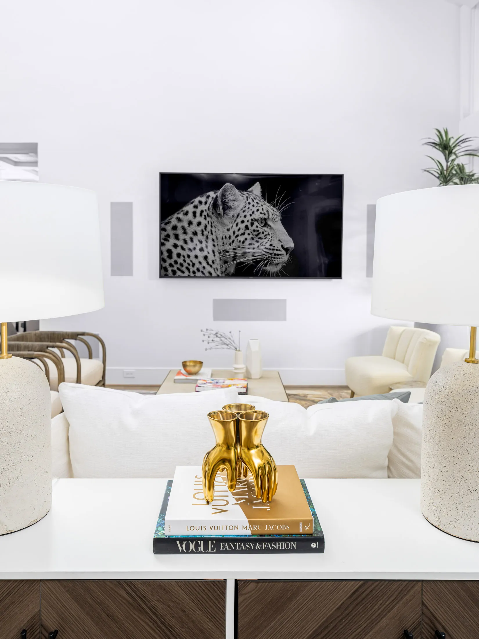 A white console table behind a sofa, decorated with fashion books and a gold hand sculpture, facing a TV displaying a black-and-white leopard image.
