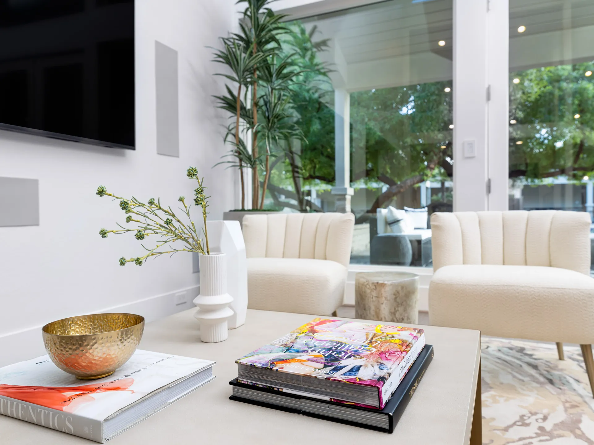 A close-up of a coffee table with gold accents, decorative books, and white vases, set before two cream chairs near large glass doors.