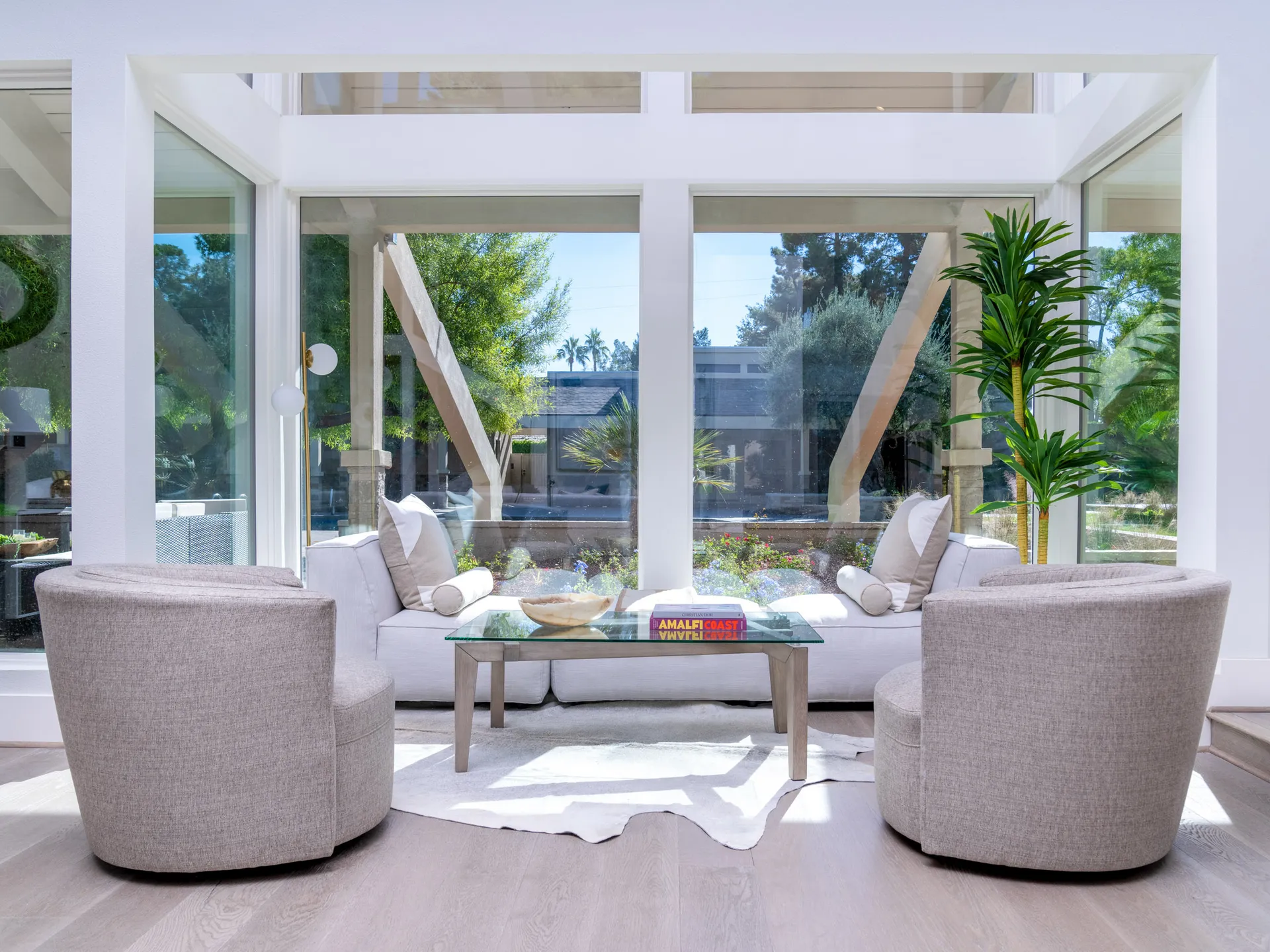 A bright corner seating area with large glass walls, two circular chairs, and a glass coffee table decorated with books and a decorative bowl.