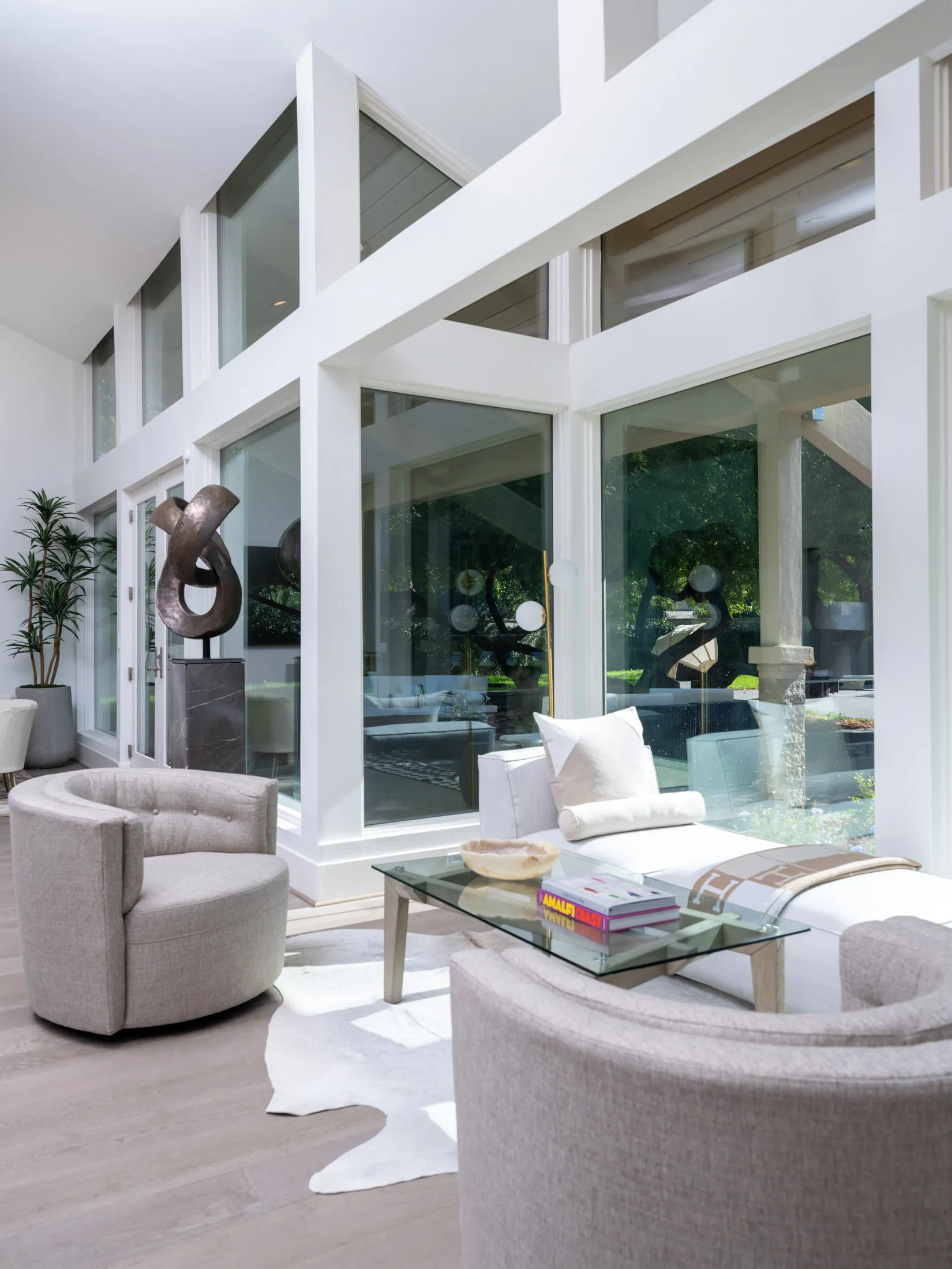 A bright seating area with floor-to-ceiling windows, two curved chairs, and a glass coffee table topped with books.