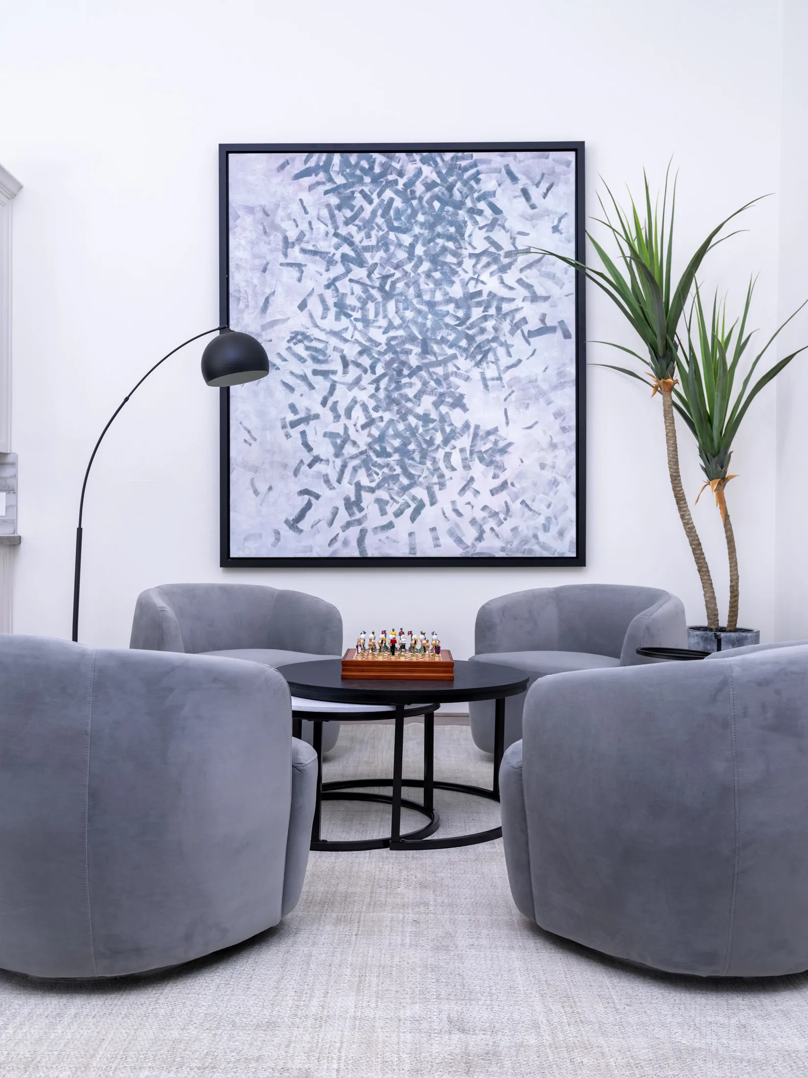 Four gray swivel chairs gathered around a black circular table with a decorative chess set, set against a large abstract painting and a tall plant.