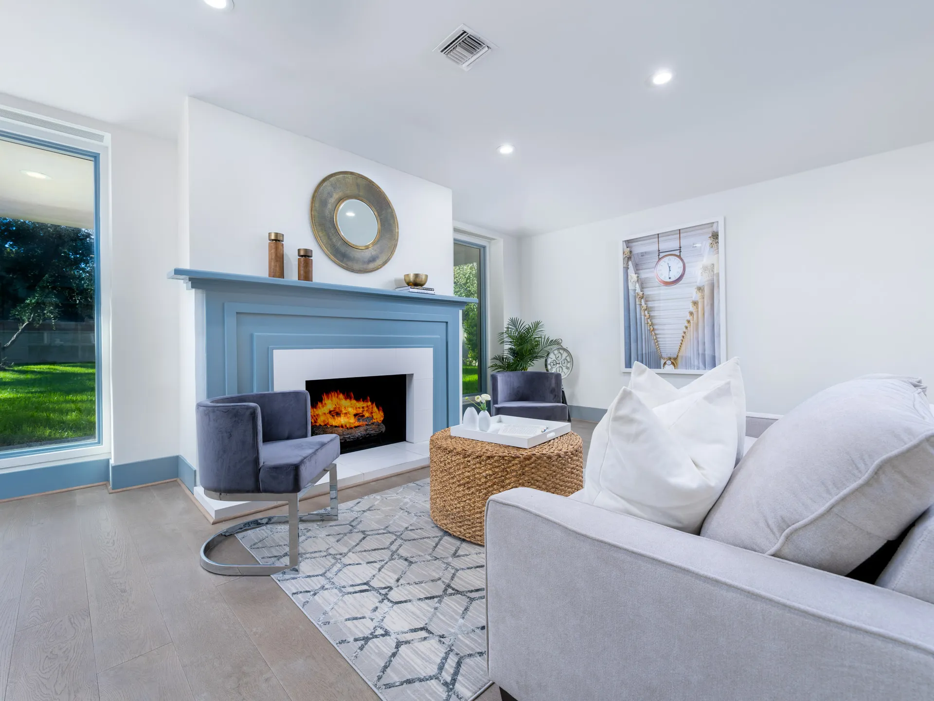 A living room featuring a painted blue fireplace with a lit fire, a gray sofa, and a velvet accent chair, framed by large windows overlooking green lawn.