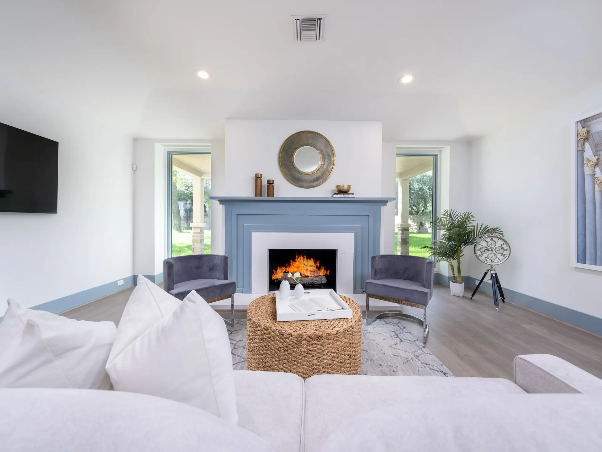 A living room showcasing a painted blue fireplace with a warm fire, two gray velvet chairs, and a woven ottoman, framed by large windows.