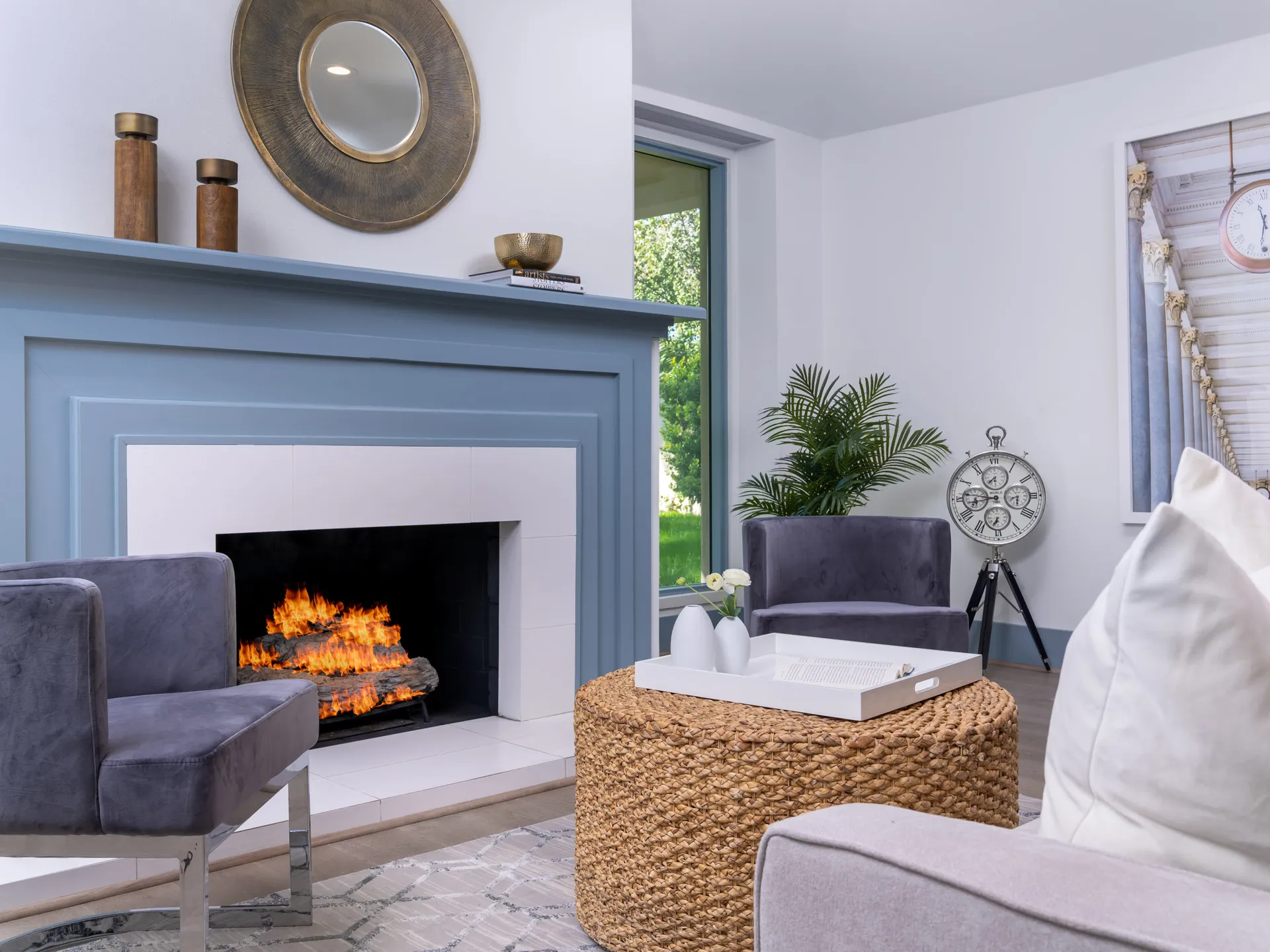 A living area with a blue-painted fireplace emitting flames, two gray chairs, and a woven ottoman topped with a white tray.