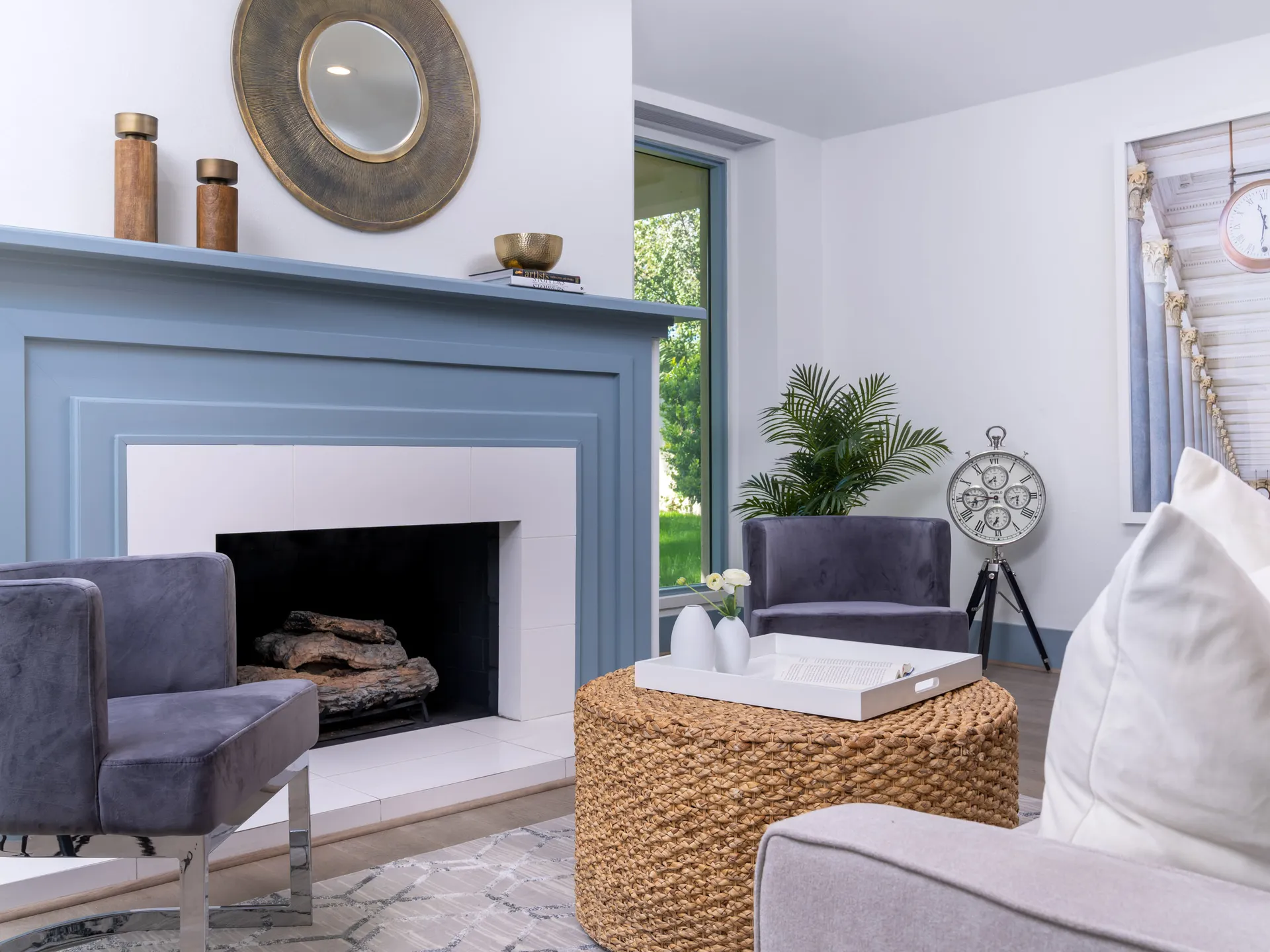 A living area with a blue-painted fireplace, two gray chairs, and a woven ottoman.