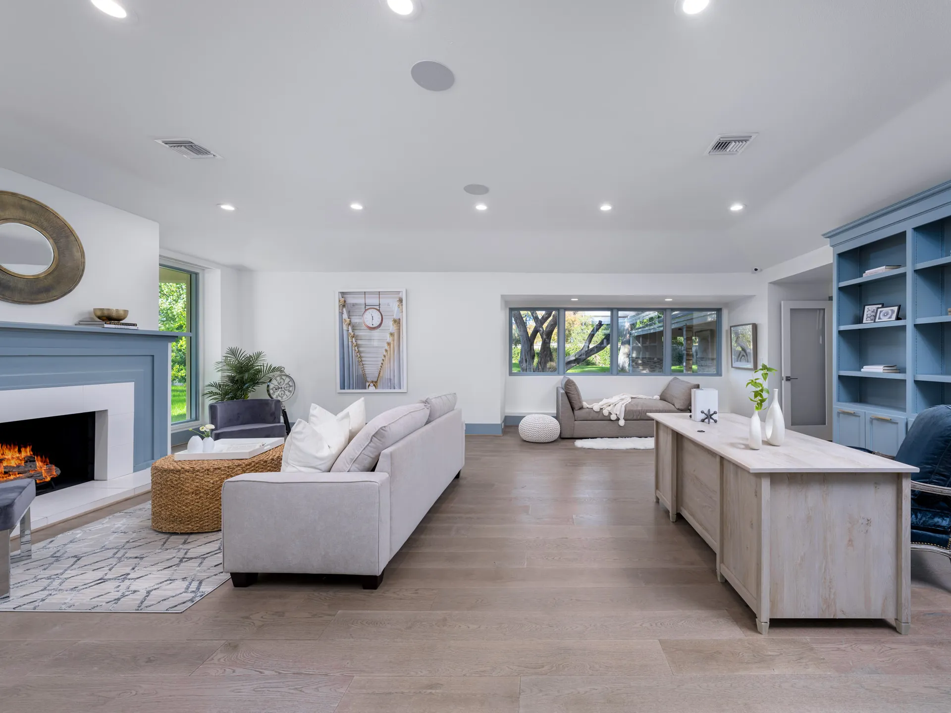 A large, open room featuring a cozy lit fireplace, a neutral sofa, and built-in blue shelves, with a desk and additional seating by expansive windows.