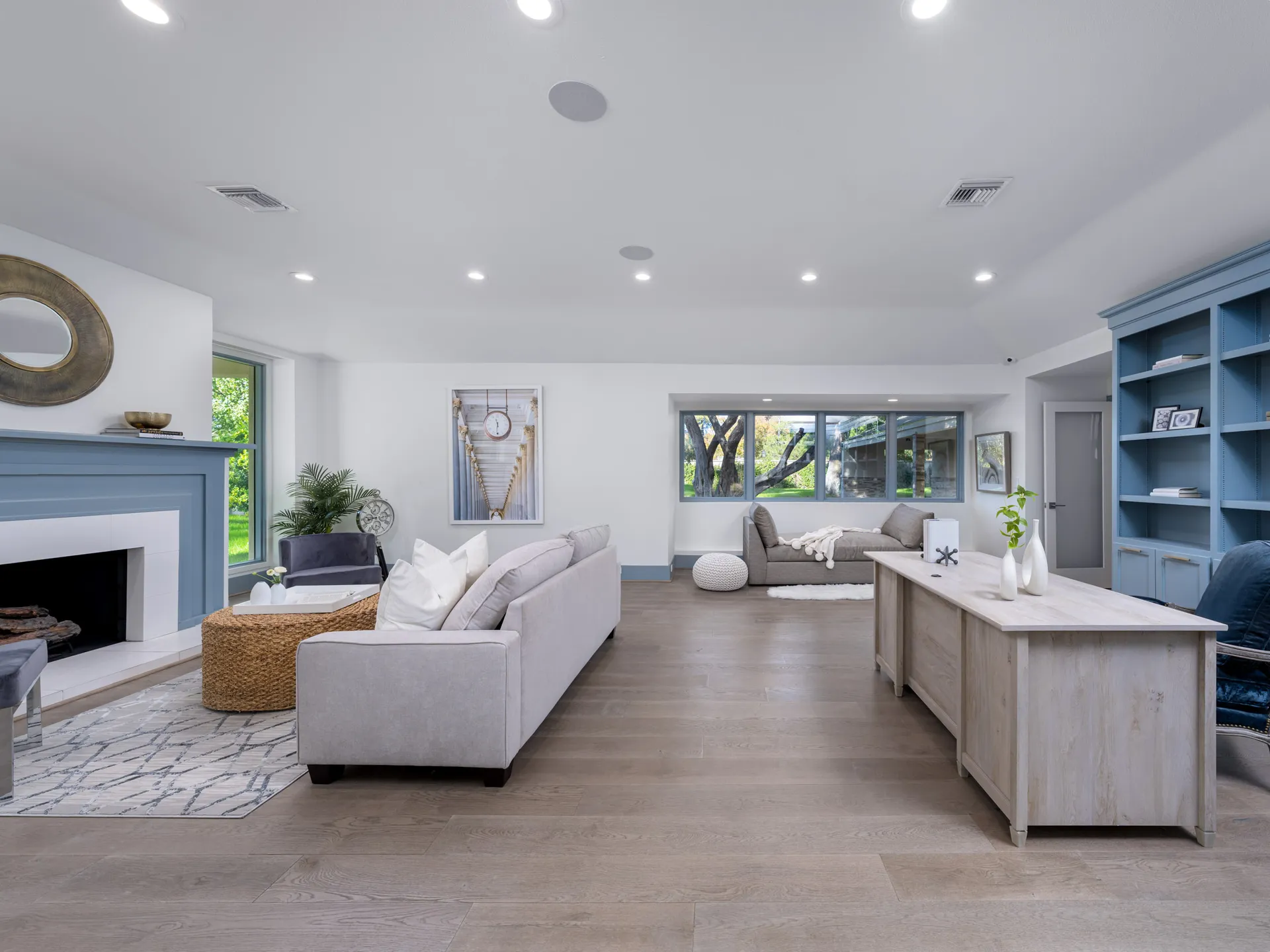 A large living area with a blue-painted fireplace, built-in shelves, and a light wood desk, featuring multiple seating areas by large windows.