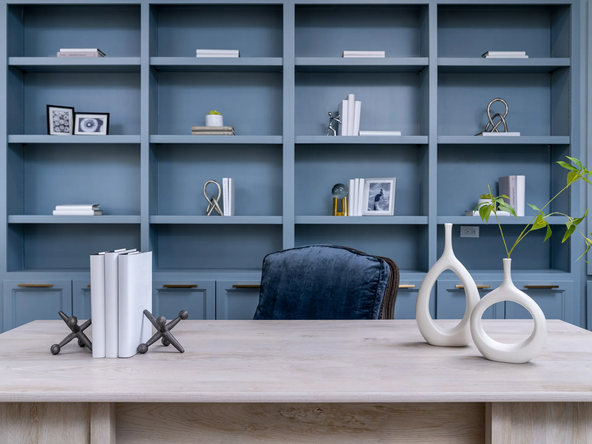 A home office desk with white sculptural vases and a plush navy chair, set against tall, painted-blue shelving.