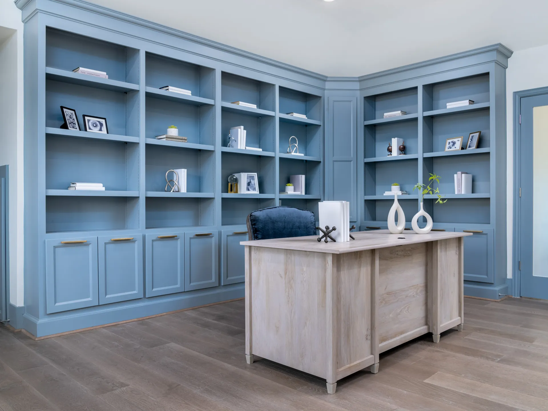 A corner home office with tall, blue built-in shelves and a light wood desk, decorated with minimal accents.
