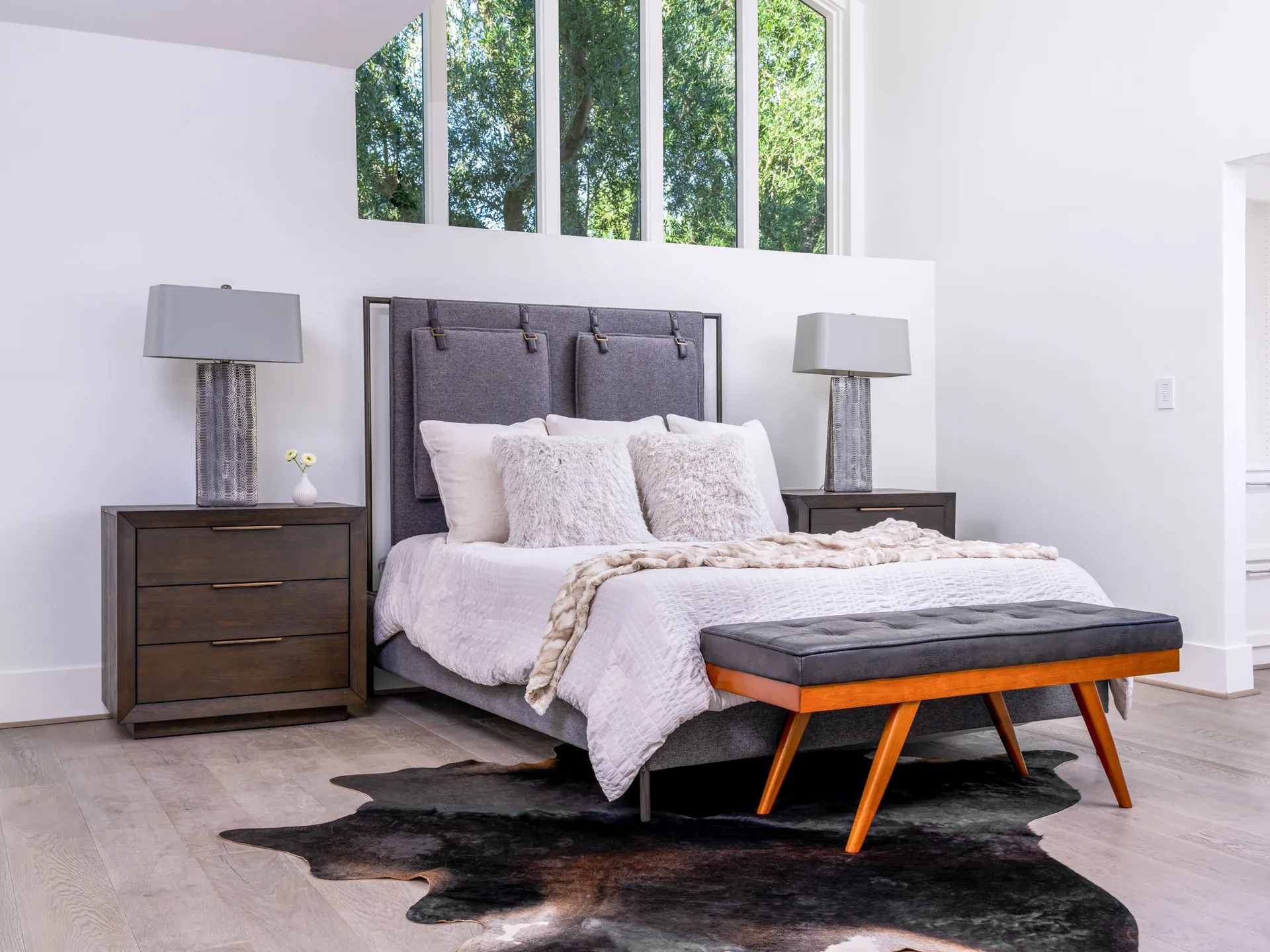 A gray upholstered bed with white and fluffy pillows, framed by large windows. A mid-century style bench sits at the foot of the bed on a cowhide rug.
