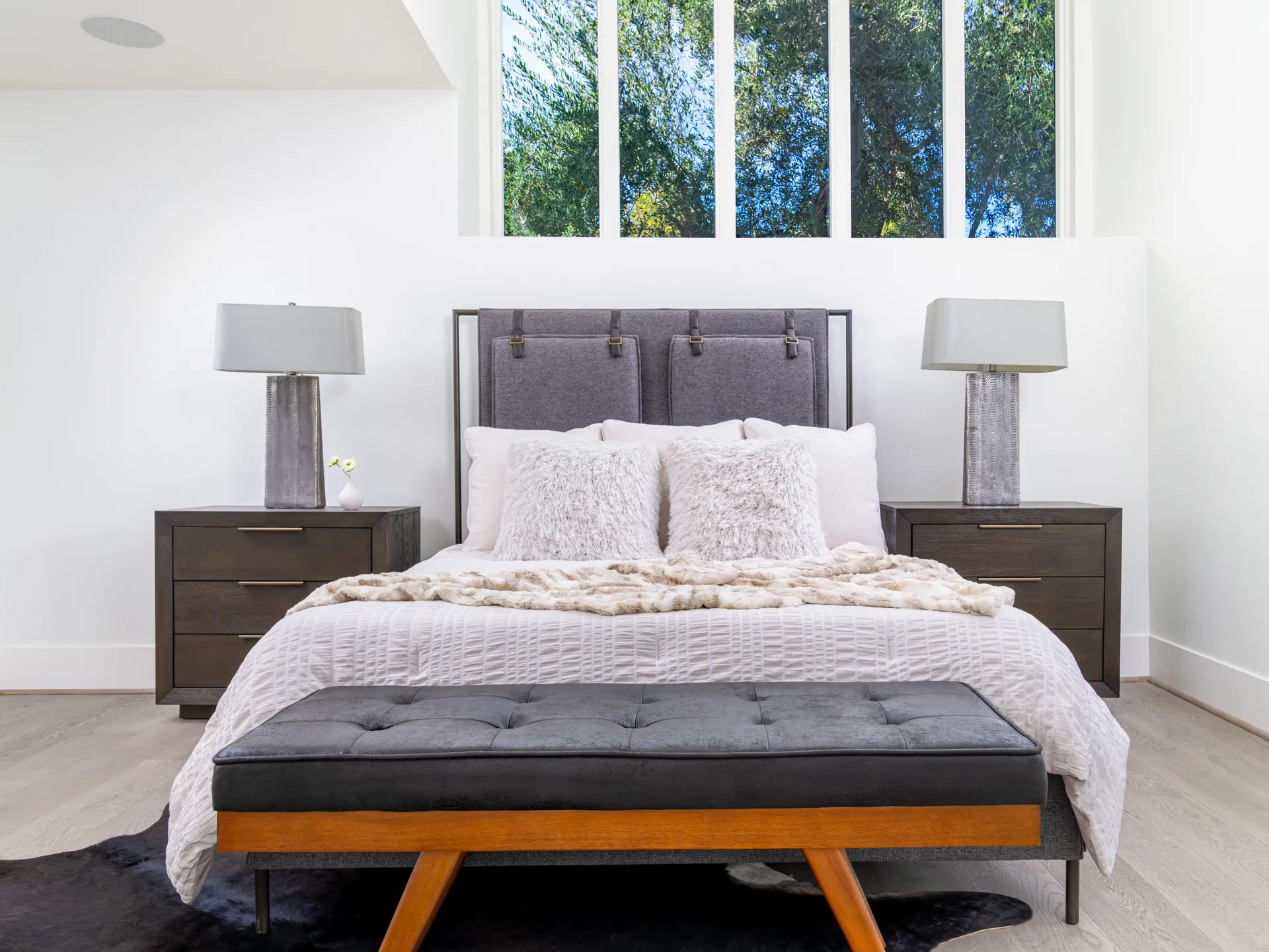 A tall bedroom with a gray upholstered headboard, white textured bedding, and a tufted bench at the foot of the bed beneath large windows.
