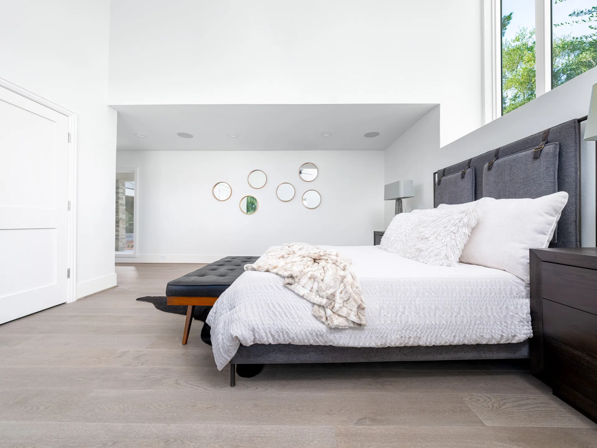 A tall, airy bedroom featuring a gray upholstered headboard, white bedding, and round mirrors on the wall.