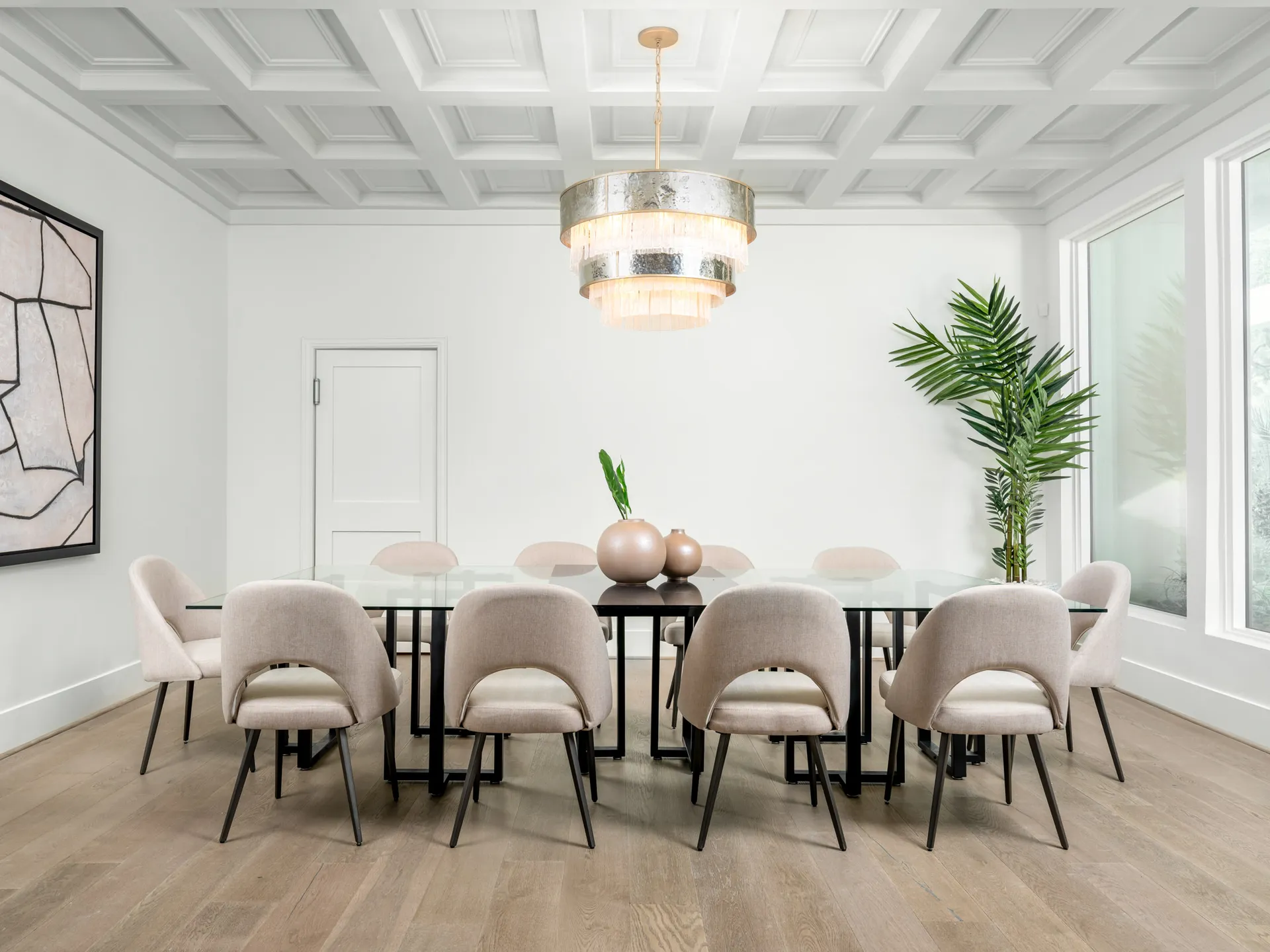 A spacious dining room with a glass-top table, curved upholstered chairs, and a tiered chandelier under a white coffered ceiling.