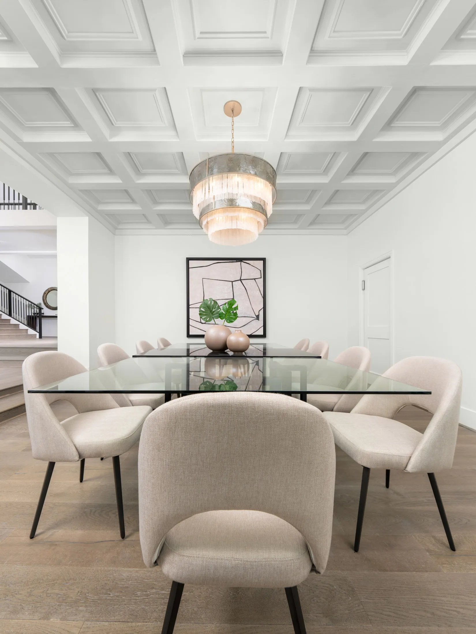 A glass-top dining table surrounded by neutral upholstered chairs beneath a tiered chandelier and a coffered ceiling.