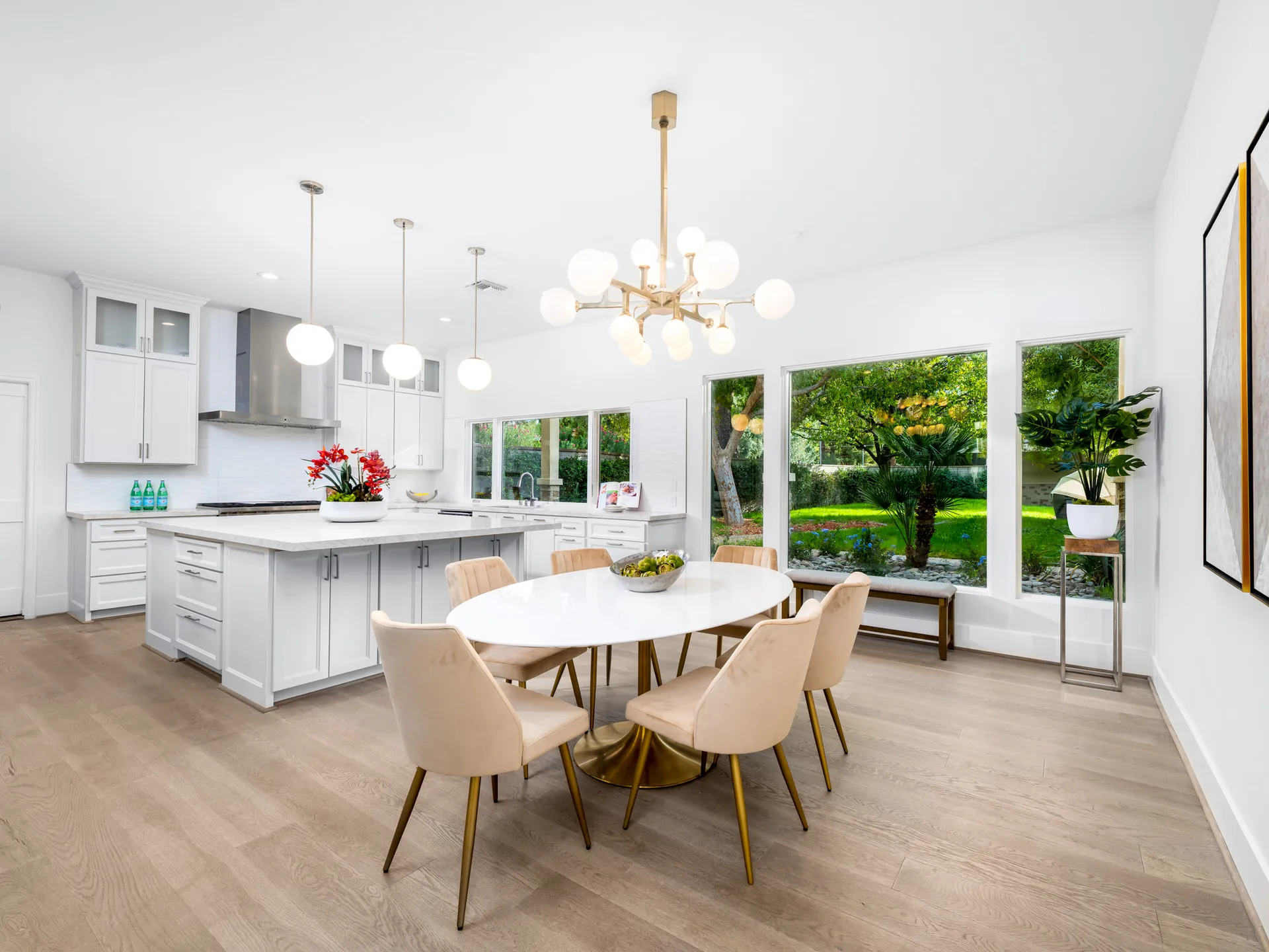 A bright kitchen with white cabinets, a large island, and a round dining table with velvet chairs near windows overlooking greenery.