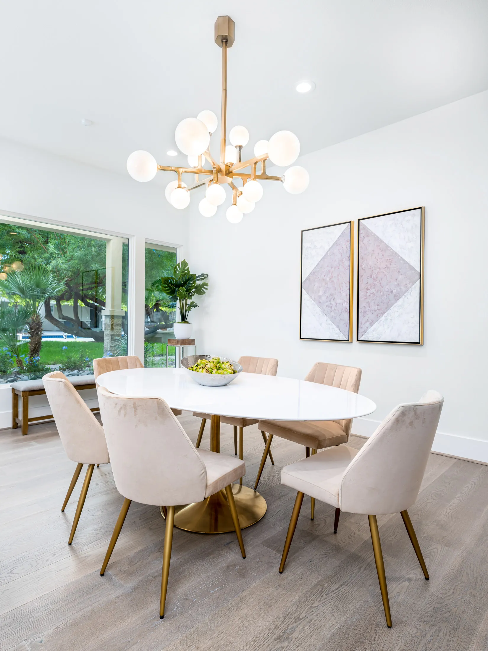 A bright dining area featuring a white oval table, velvet chairs with gold legs, and a sculptural brass chandelier.