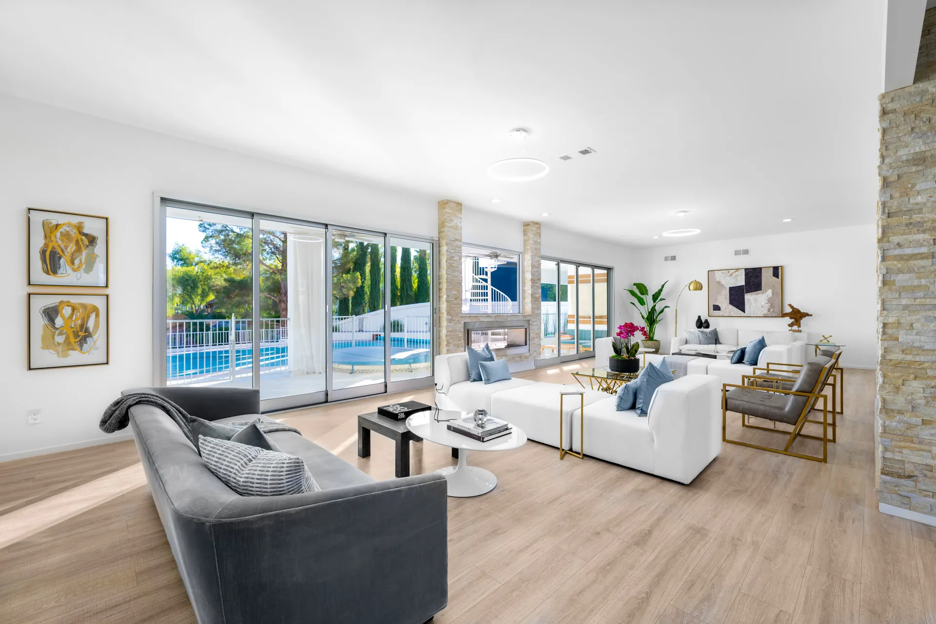 A white living room with large sliding doors opening to a pool, a stone fireplace, gold accent chairs, and plush seating.