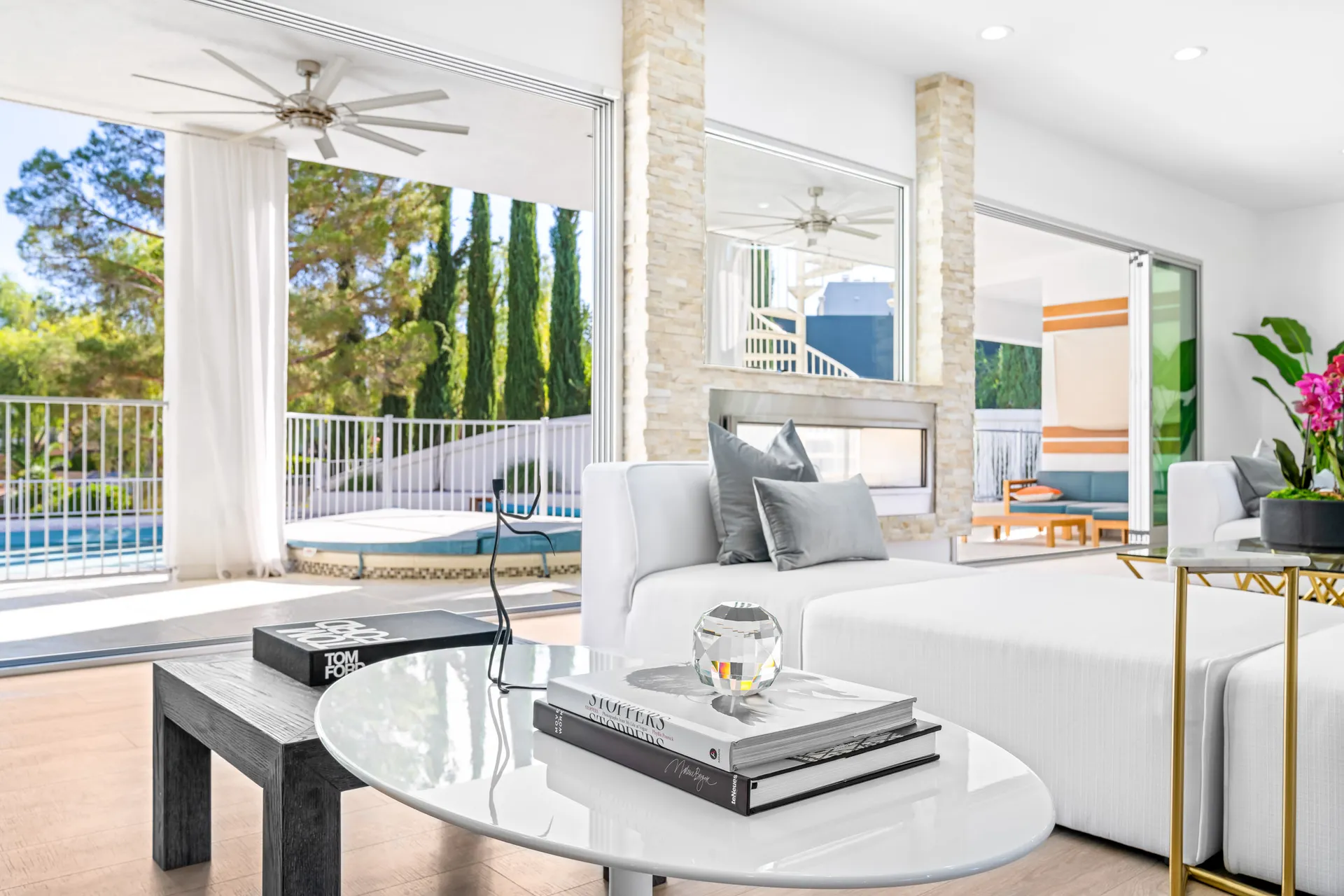 A white sofa with gray pillows in a bright living area, facing a stone fireplace and large sliding doors that open to a pool and spa.