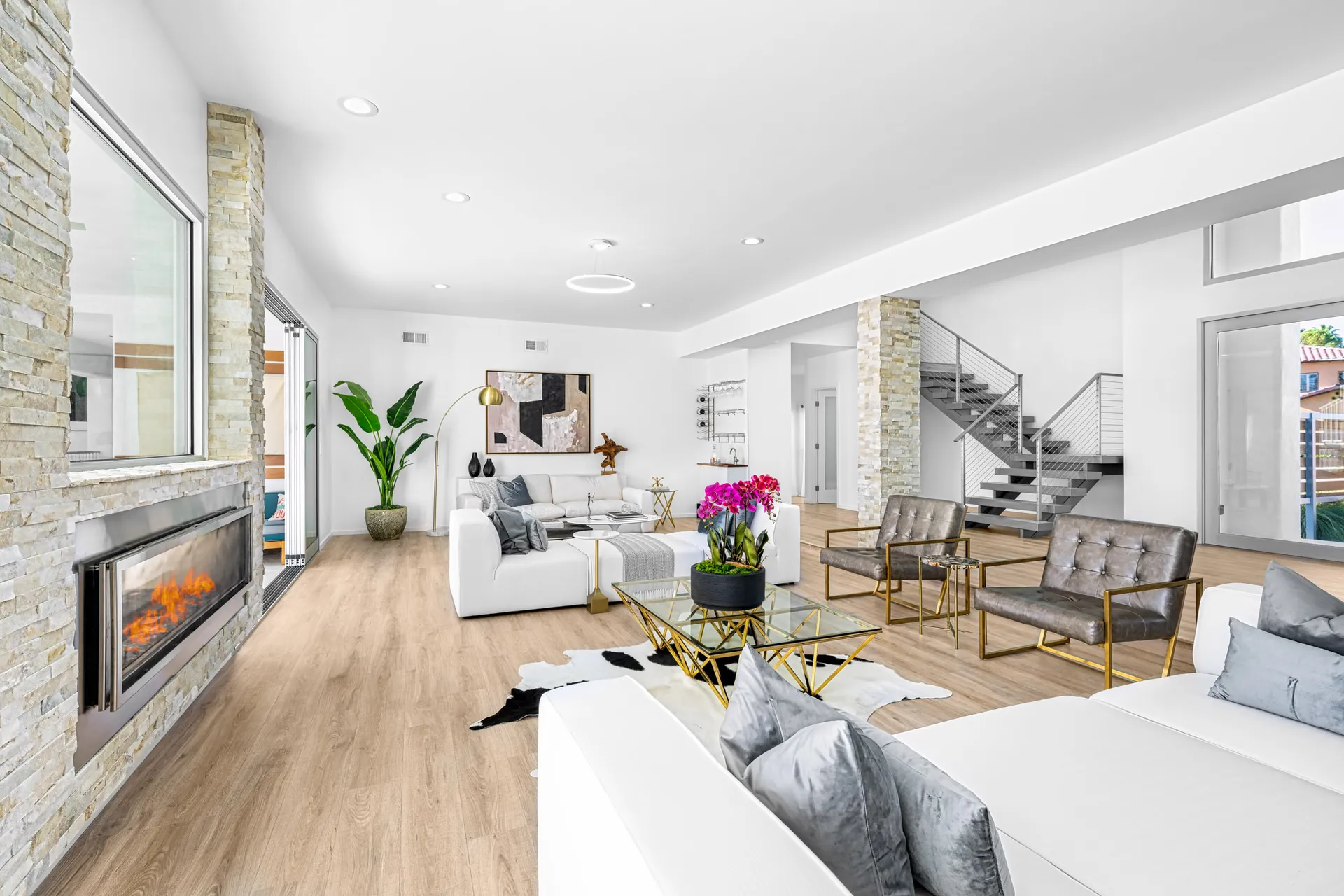 A bright living room featuring a white sectional, a stone-accent fireplace, a gold coffee table with pink orchids, and a modern metal staircase.
