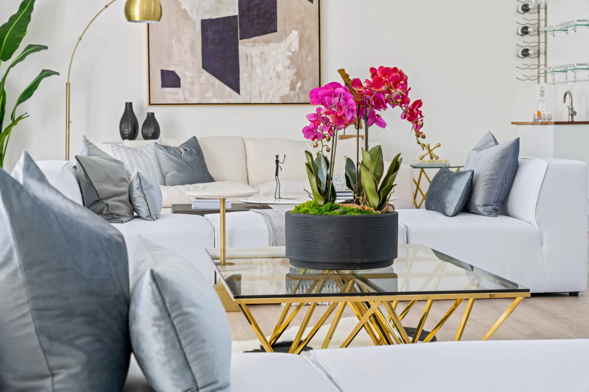A white seating arrangement with pink orchids on a gold coffee table in a bright living space.