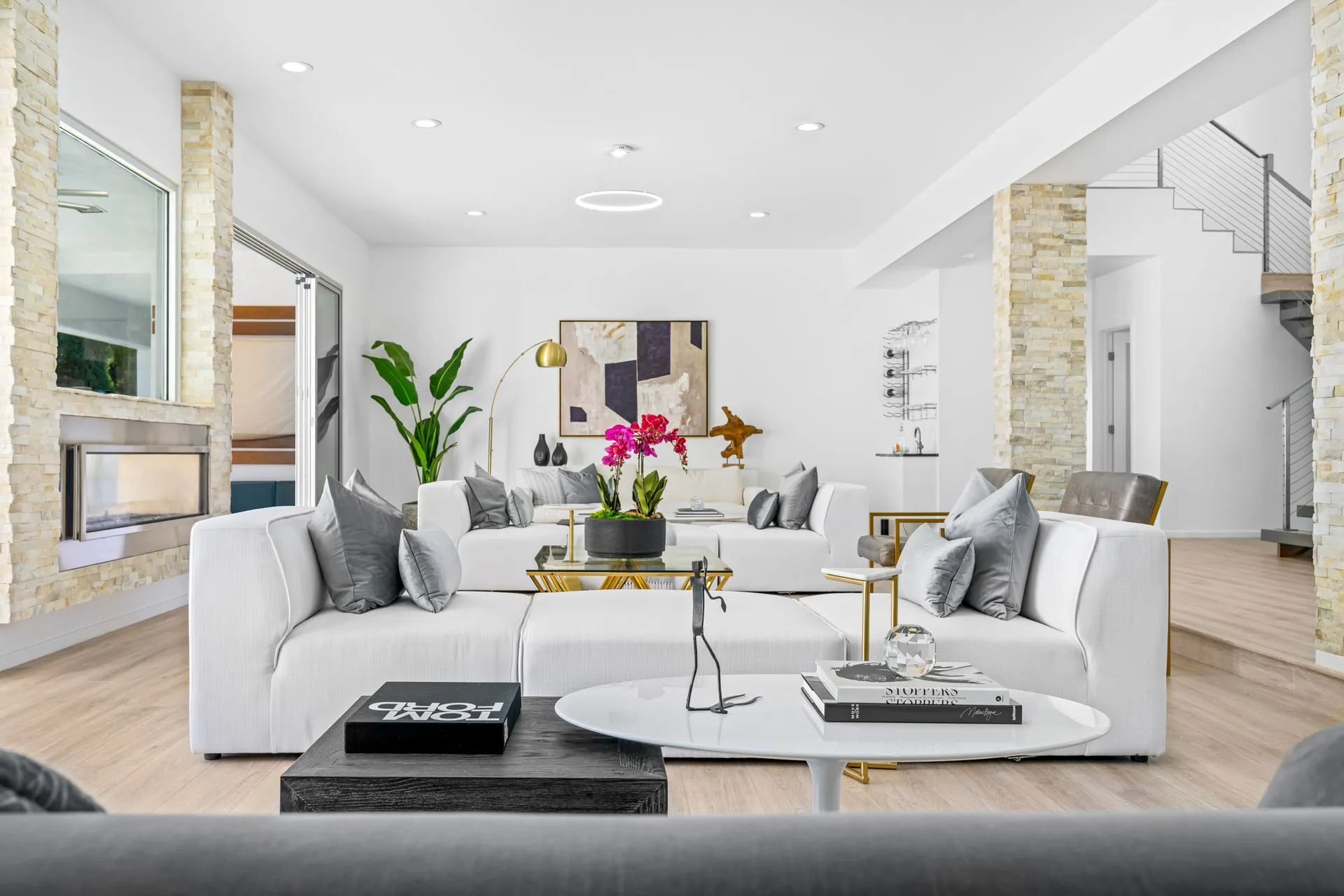 A light-filled living room with white sofas, a pink orchid centerpiece, stone accents, and a modern staircase in the background.