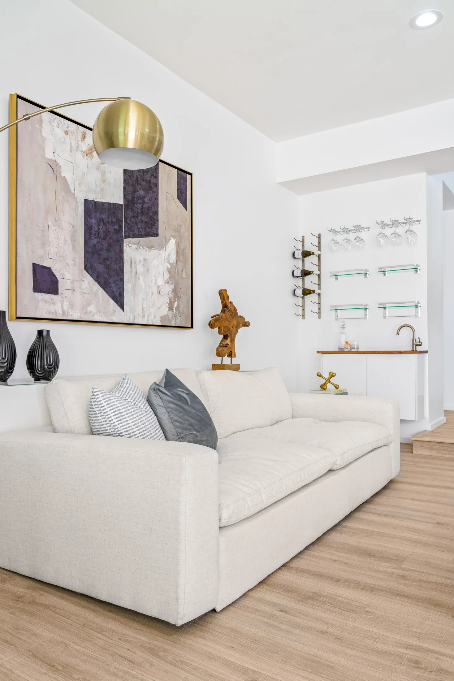 A white sofa with accent pillows sits beneath an abstract painting, with a small bar and hanging glass racks nearby.