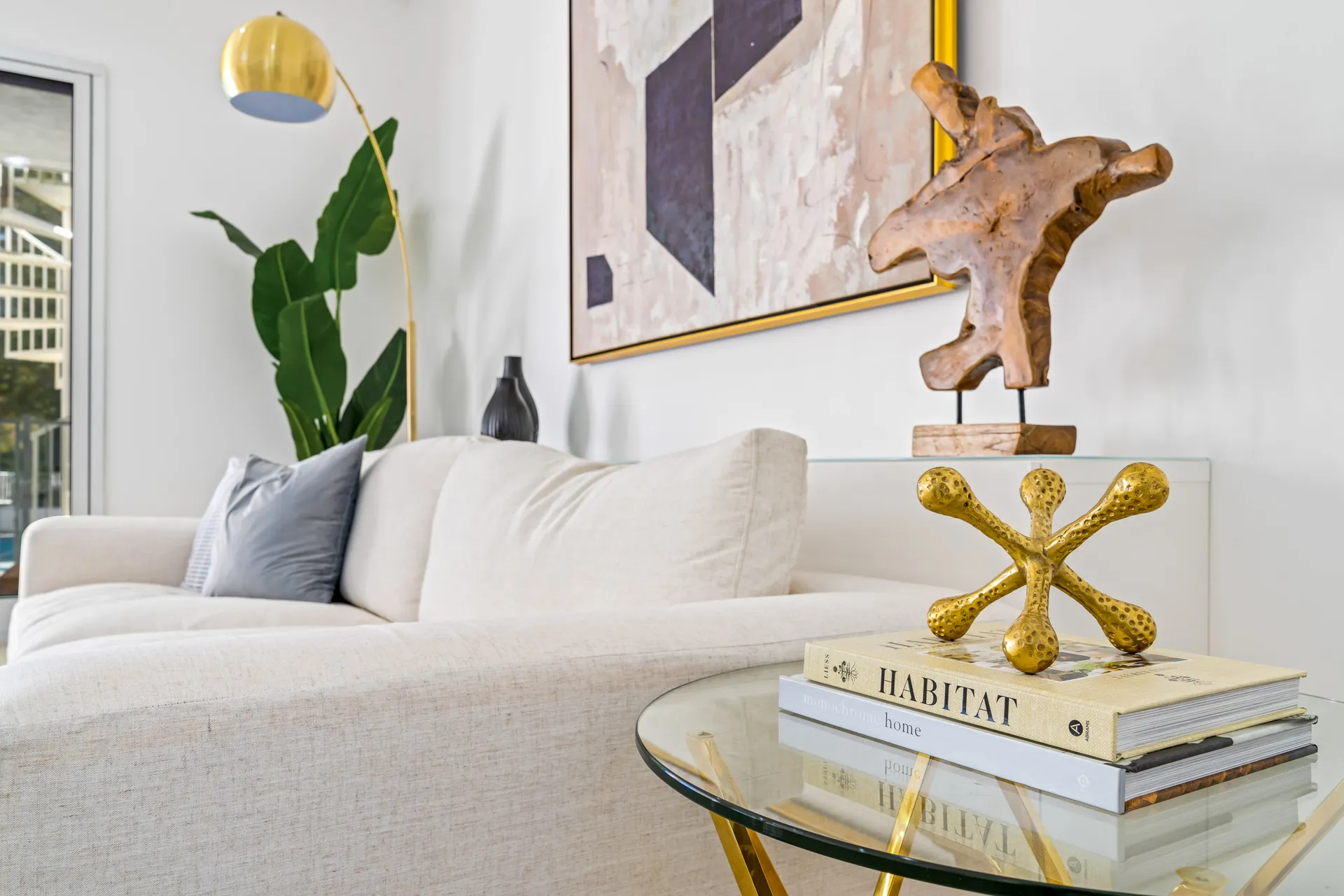 A white sofa with gray pillows, a gold floor lamp, and abstract art on the wall. A small glass table holds decorative books and a gold sculpture.
