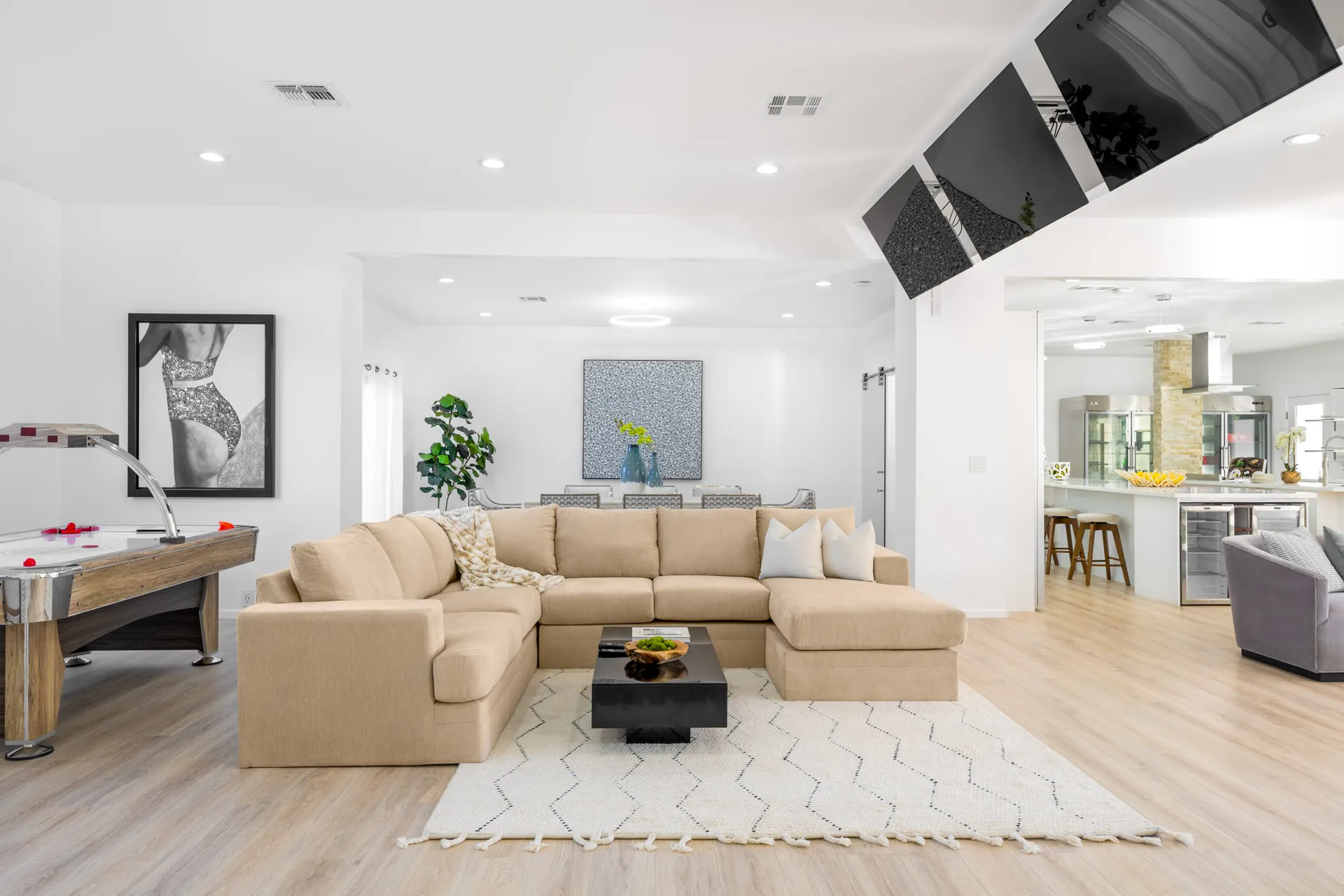 A bright open living area with a beige sectional, black coffee table, an air hockey table, and wood flooring.